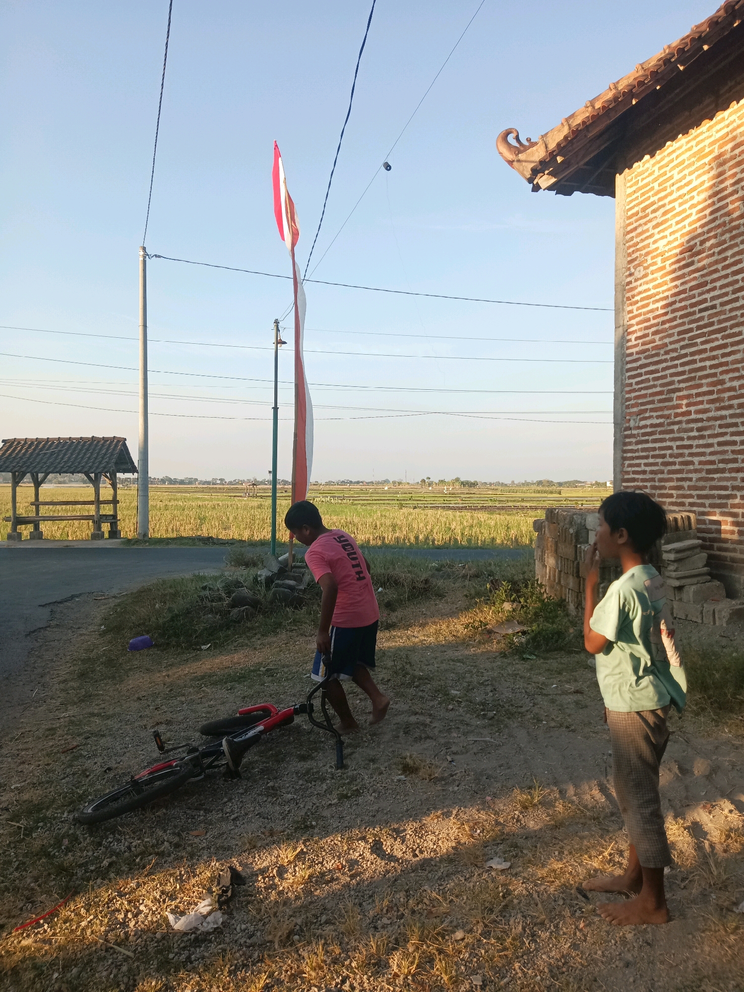 In the city of Gondang, Indonesia, a street scene unfolds under a clear sky. Two boys are present; one, wearing a pink t-shirt and blue shorts, stands near a fallen red bicycle. The other boy, in a light blue t-shirt and checkered trousers, stands with his back to the viewer. The environment includes a brick building with a distinctive tiled roof on the right, a road, a grassy area, and a distant view of a rice field. A tall pole with an Indonesian flag is visible in the background. The lighting suggests it is late afternoon, casting long shadows.