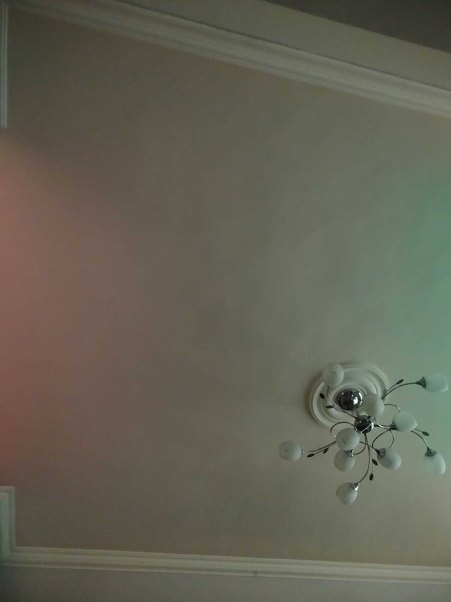 The image shows a low-angle shot of a room's ceiling. The ceiling is off-white, with decorative molding along its edges. A modern chandelier is mounted in the lower right portion of the frame. It features multiple small, rounded glass shades attached to curving metal arms, centered around a circular decorative base. The lighting conditions are dim, with red tint on the left and green tint on the right. The location is likely a residential or commercial space, but there are no clues to indicate Yerevan, Armenia.