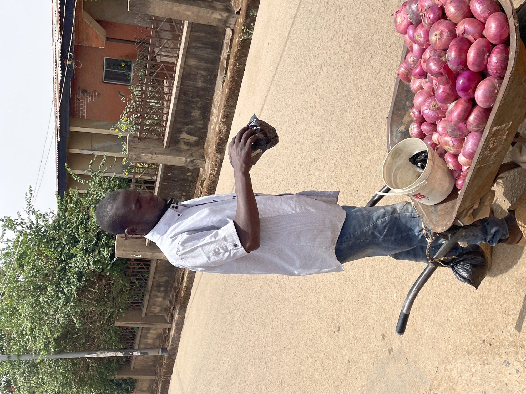A man stands on a paved road in Jos, Nigeria, beside a wheelbarrow overflowing with red onions. He is wearing a white collared shirt and blue jeans. He holds a small black plastic bag in his left hand. A white bucket sits in the wheelbarrow among the onions, also containing a black plastic bag. A black plastic bag is tied to the wheelbarrow handle. Buildings and vegetation are visible in the background. The lighting suggests it is daytime.