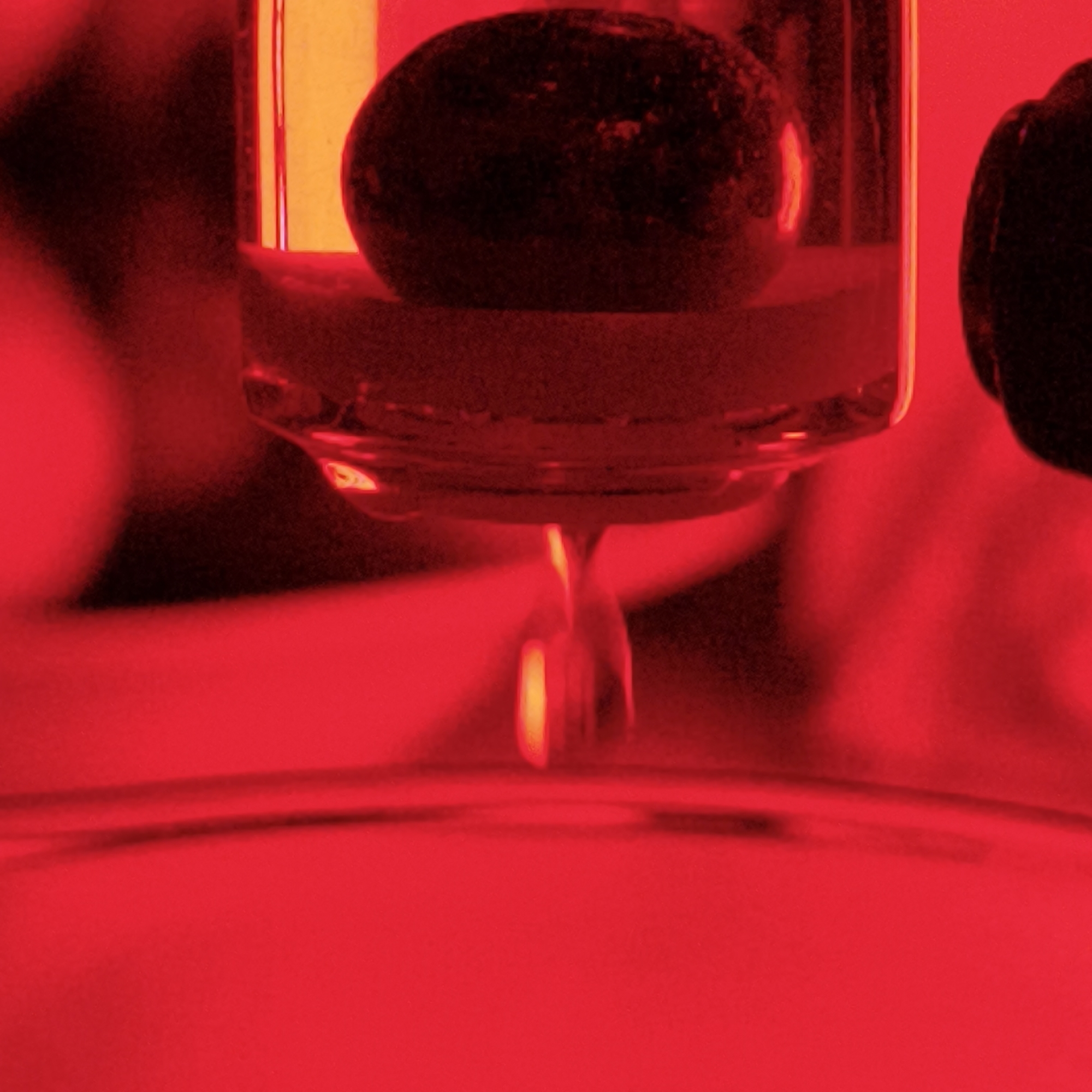 A close-up image, bathed entirely in red light, captures a laboratory or scientific setup. A clear, cylindrical glass container holds a dark, spherical object submerged in a liquid. A droplet of liquid is in the process of detaching from the bottom of the container, heading towards what appears to be a dish or another container below. The illumination does not allow for precise identification of the liquid, but it seems viscous. The cylindrical container is partially obscured, suggesting it might be attached to other equipment. The location in Парэчча, Belarus, is not discernible from the provided visual information.