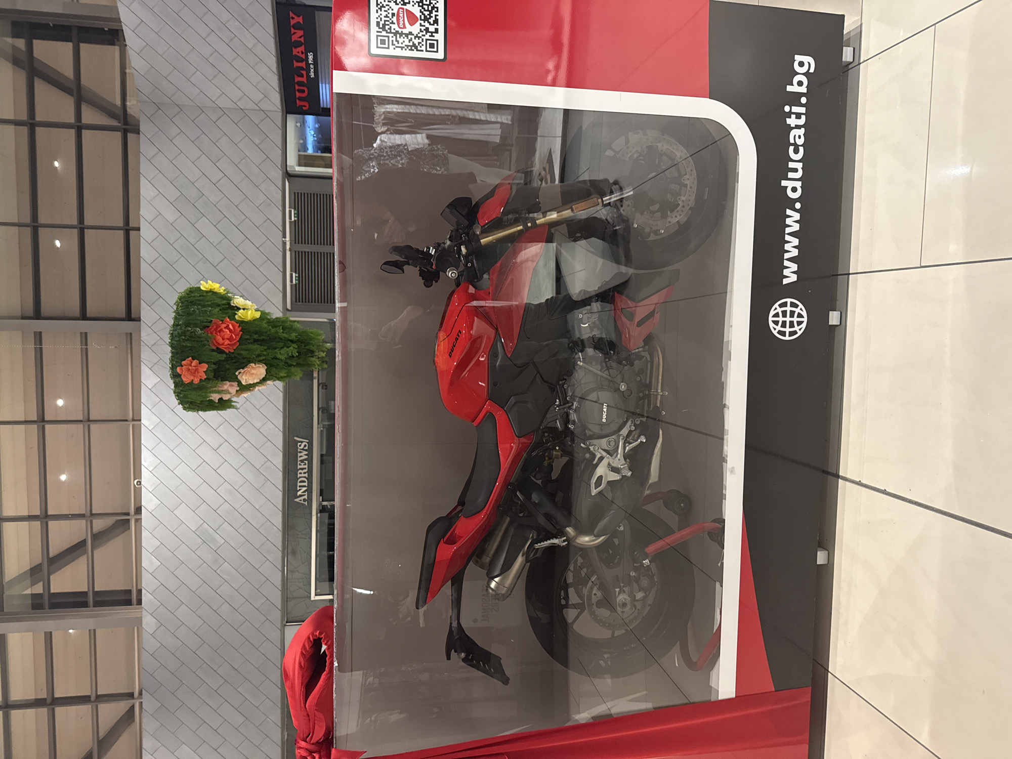 The image shows a red Ducati motorcycle displayed in a transparent enclosure. A sign on the enclosure reads "www.ducati.bg". The background includes a view of an interior space with a building facade featuring a store sign that reads "ANDREWS/" and "JULIANY since 1985". A decorative floral arrangement is next to the enclosure. Based on the signage and the .bg domain, the location is likely Sofia, Bulgaria. No people are visible in the scene. The motorcycle is the central object, presented as part of a promotional display.