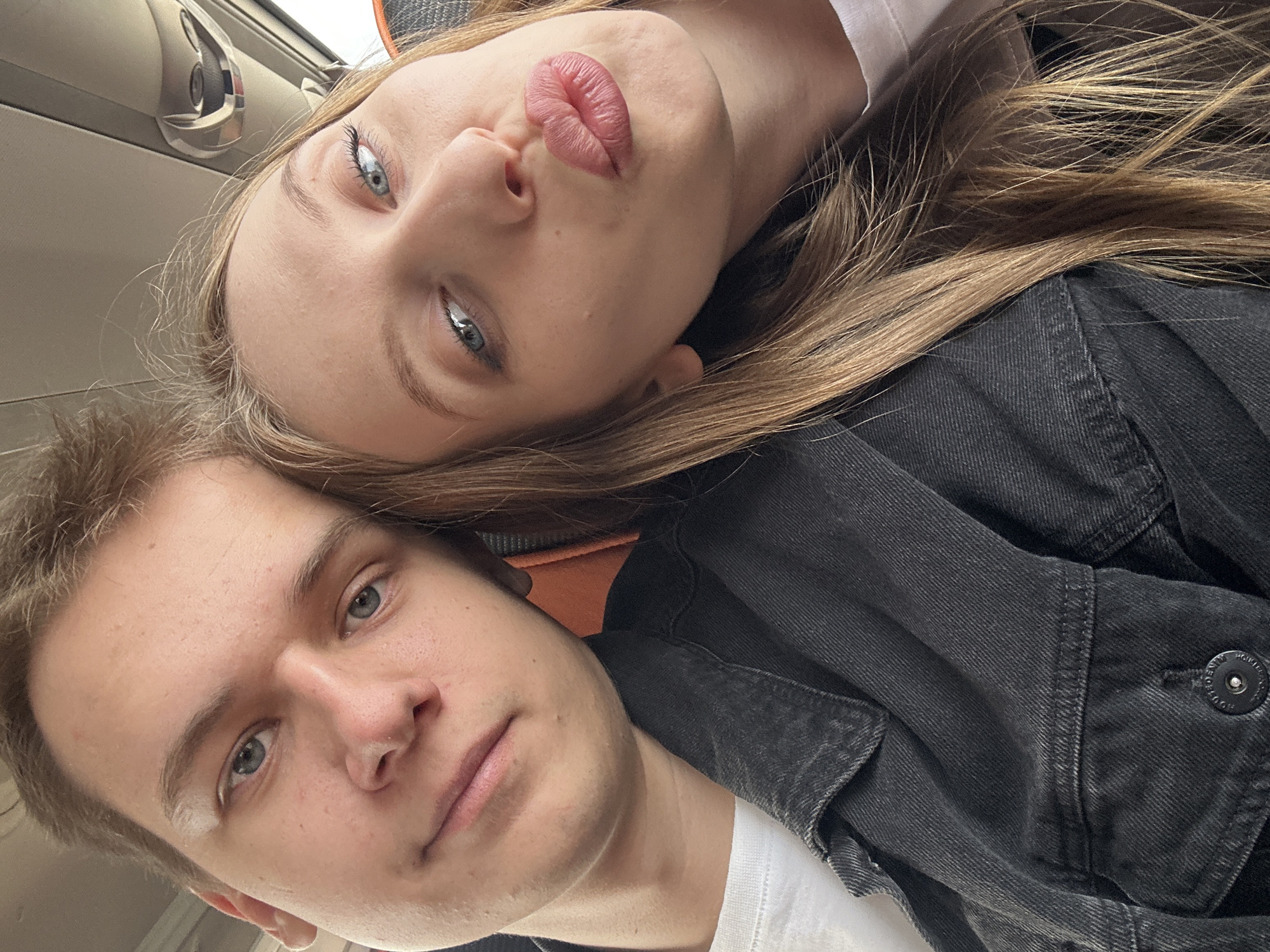 A selfie captures two fair-skinned individuals, a male and a female. The woman is positioned above the man, her head tilted. She has long, light-brown hair and blue eyes. Her lips are pursed. The man has short, light-brown hair and blue eyes. Both are wearing white shirts, and the woman is wearing a black jacket. The photo seems to be taken in a vehicle. Location cannot be determined from the image.