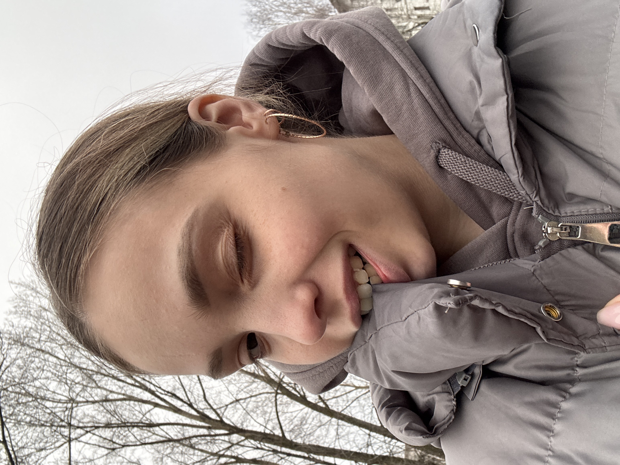 The image is a selfie taken by a young woman outdoors. She is wearing a grey hooded sweatshirt under a grey puffer jacket. Her hair is light brown and she is wearing a gold hoop earring. She is biting on the collar of her jacket. Bare trees are visible in the background. It is impossible to determine from the image if the location is Chaikovsky, Russia.