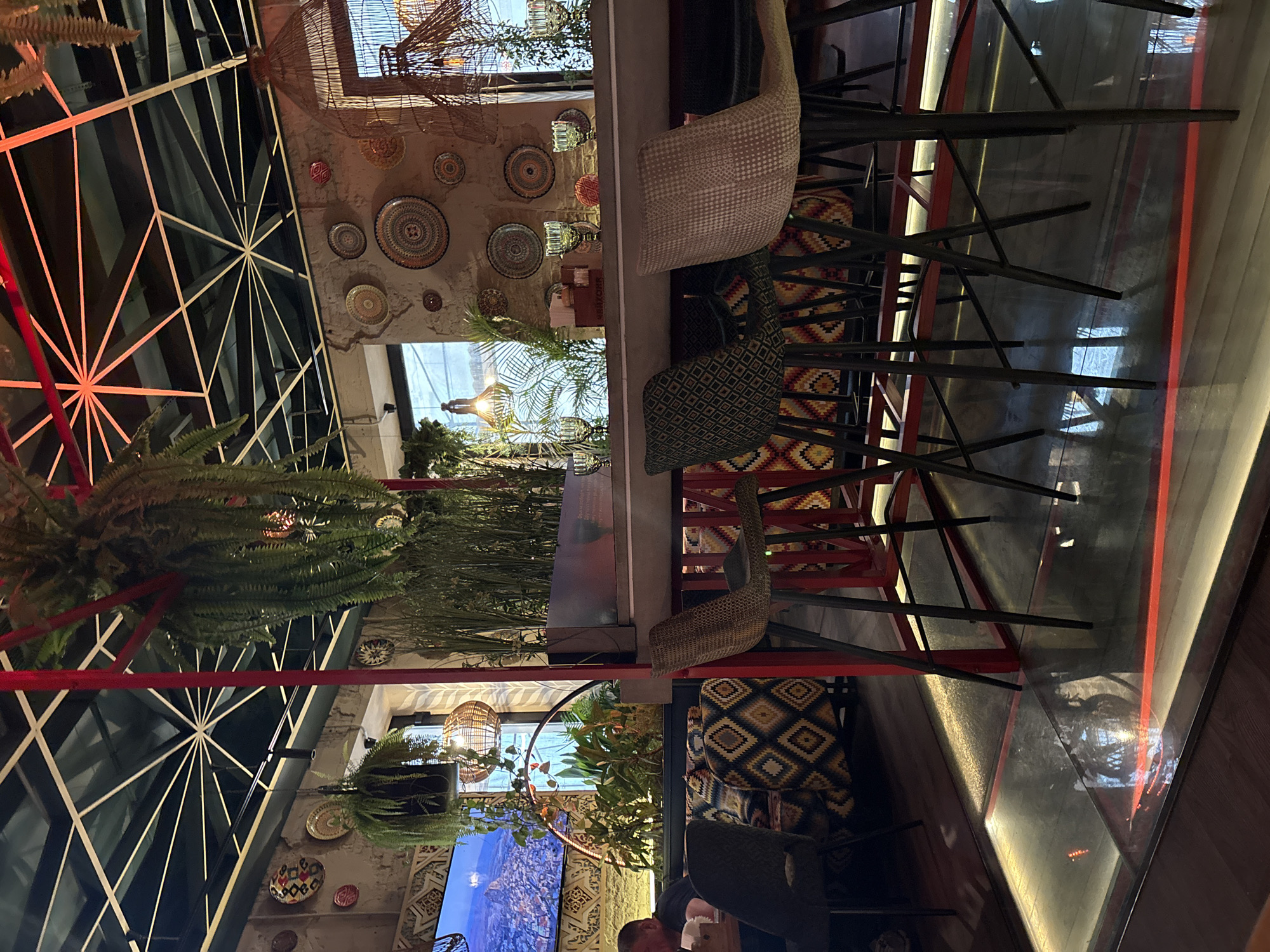 The image shows the interior of a bar or restaurant, potentially located in Saint Petersburg, Russia, based on architectural and decorative elements often found in that city. The space features a high ceiling with an elaborate, geometric metal framework and hanging plants. The walls are adorned with decorative plates. A long bar is visible, lined with high chairs with patterned cushions. A person is seated at one of the tables. The overall atmosphere suggests a trendy and modern establishment.