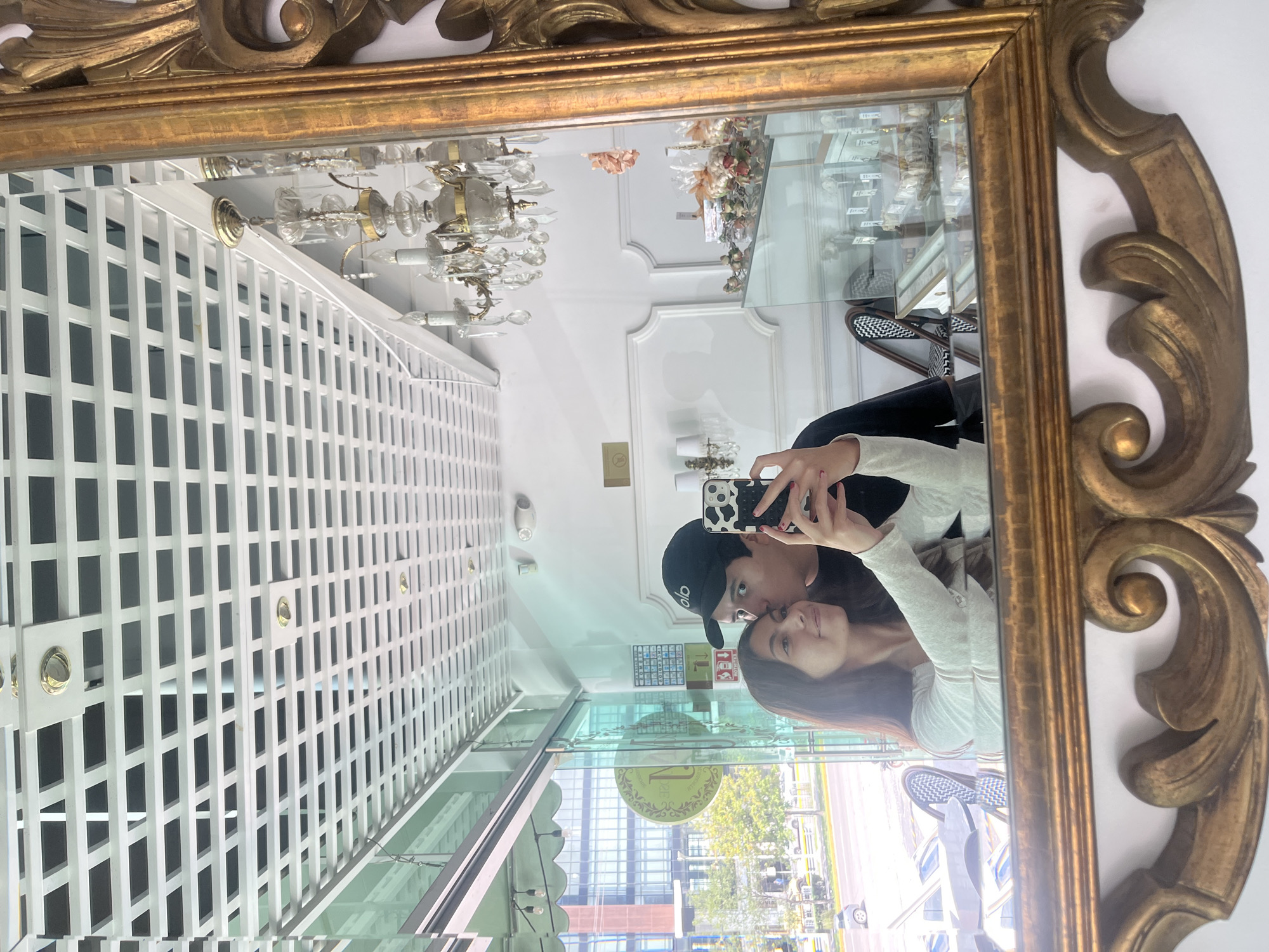 A young couple is taking a mirror selfie in what appears to be a shop. The man, wearing a black baseball cap and a dark t-shirt, is kissing the woman on the cheek. The woman, with long brown hair, is wearing a light-colored long-sleeved shirt. She is holding a smartphone with a cow-print case, taking the picture.
The mirror is ornate with a gold, carved frame. Behind the couple, the reflection shows a white ceiling with a white lattice design, a crystal chandelier, and a display case filled with items that appear to be gifts or confectioneries. The shop has large glass windows, through which a street scene is visible, including a building with multiple floors and trees. The location is identified as San Bernardino Tlaxcalancingo, Mexico.