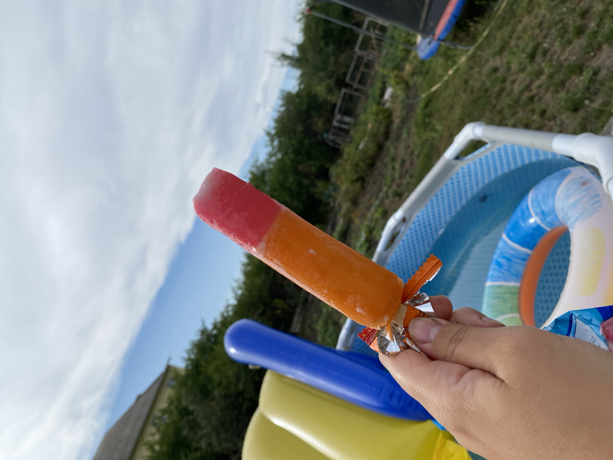 The media file shows a hand holding a red and orange popsicle in front of a blurred background. The background consists of a backyard scene with a blue inflatable pool, some greenery, and a trampoline. The sky is visible in the upper part of the image, with some clouds. The overall setting appears to be a sunny day, possibly in the summer, given the presence of the pool and popsicle. The image is shot from a low angle, making the popsicle the main subject. There are no discernible people visible, only the hand holding the popsicle. The mood appears to be casual and summery. There is no visible text in the image. The location is Kropyvnytskyi, Ukraine.
