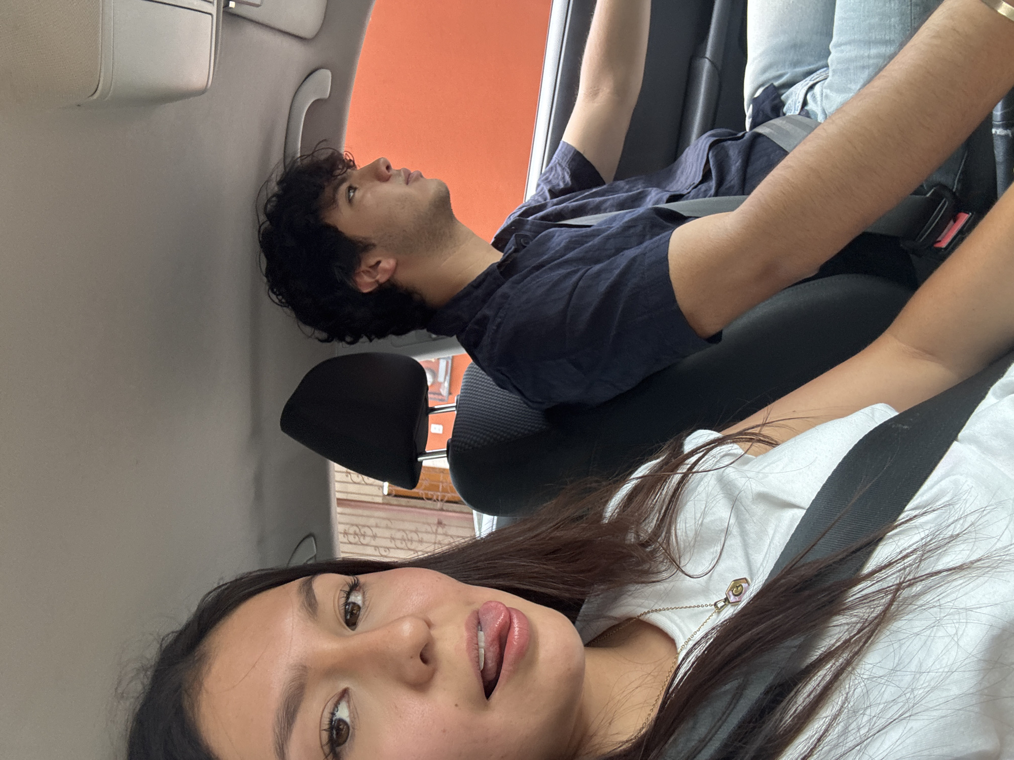Inside a car, a young woman with long brown hair and a white t-shirt is wearing a seatbelt and has her tongue slightly out. Behind her and to the left, a young man with curly dark hair, wearing a navy shirt and jeans with a seatbelt, is looking up with his arm reaching toward the car's interior. The interior of the car is primarily gray. Outside the window appears to be an orange-colored building, possibly indicating they are in the vicinity of a commercial or public building. The photo appears to be taken inside a vehicle, facing upwards at an angle, which suggests a selfie taken while inside a moving or stationary car. No specific indicators of Oaxaca City, Mexico are visible.