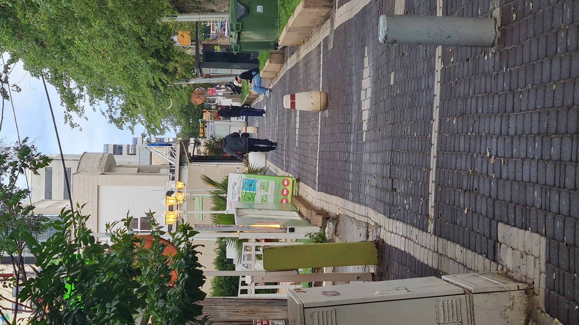 The image depicts a street scene in Nahariyya, Israel. The view is from a sidewalk paved with dark gray bricks, edged with a white painted line. The sidewalk is bordered by street furniture and greenery. A cylindrical concrete bollard stands on the right side of the sidewalk. Further down the sidewalk, three individuals are present: one bending over, one standing, and one sitting on a bench. Green trash containers are situated adjacent to the bench. On the left side of the sidewalk, there are signs and plants in front of what appears to be a commercial establishment. The architecture is a mix of low-rise buildings. Overhead, there is a partial view of the sky and the canopy of trees.