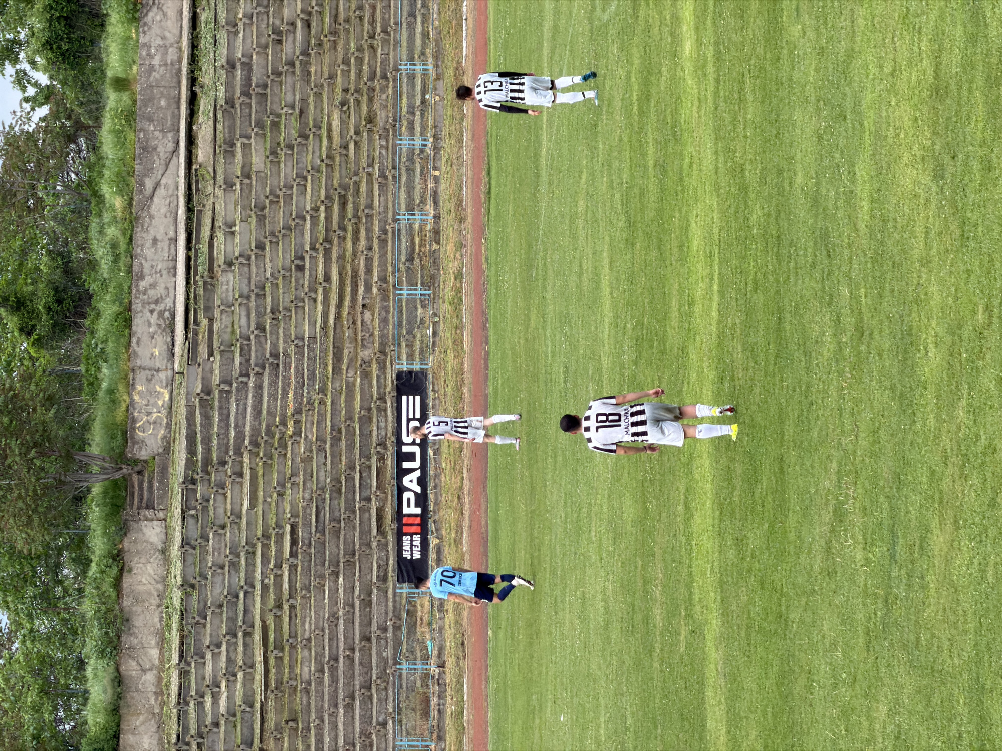 The image shows a soccer field next to an old, weathered stadium in Svishtov, Bulgaria. The stadium has concrete seating that is in disrepair. A "PAUSE" banner is attached to the stadium wall. On the field are four soccer players in uniforms. One player wears a light blue jersey with the number "70" on the back. Two players wear black and white striped jerseys with the numbers "18" and "5" respectively. The name "Malchika" is visible on the back of the player wearing number "18". The fourth player wears a similar striped jersey with the number "13" on the back. The players are positioned on the green grass of the soccer field.
