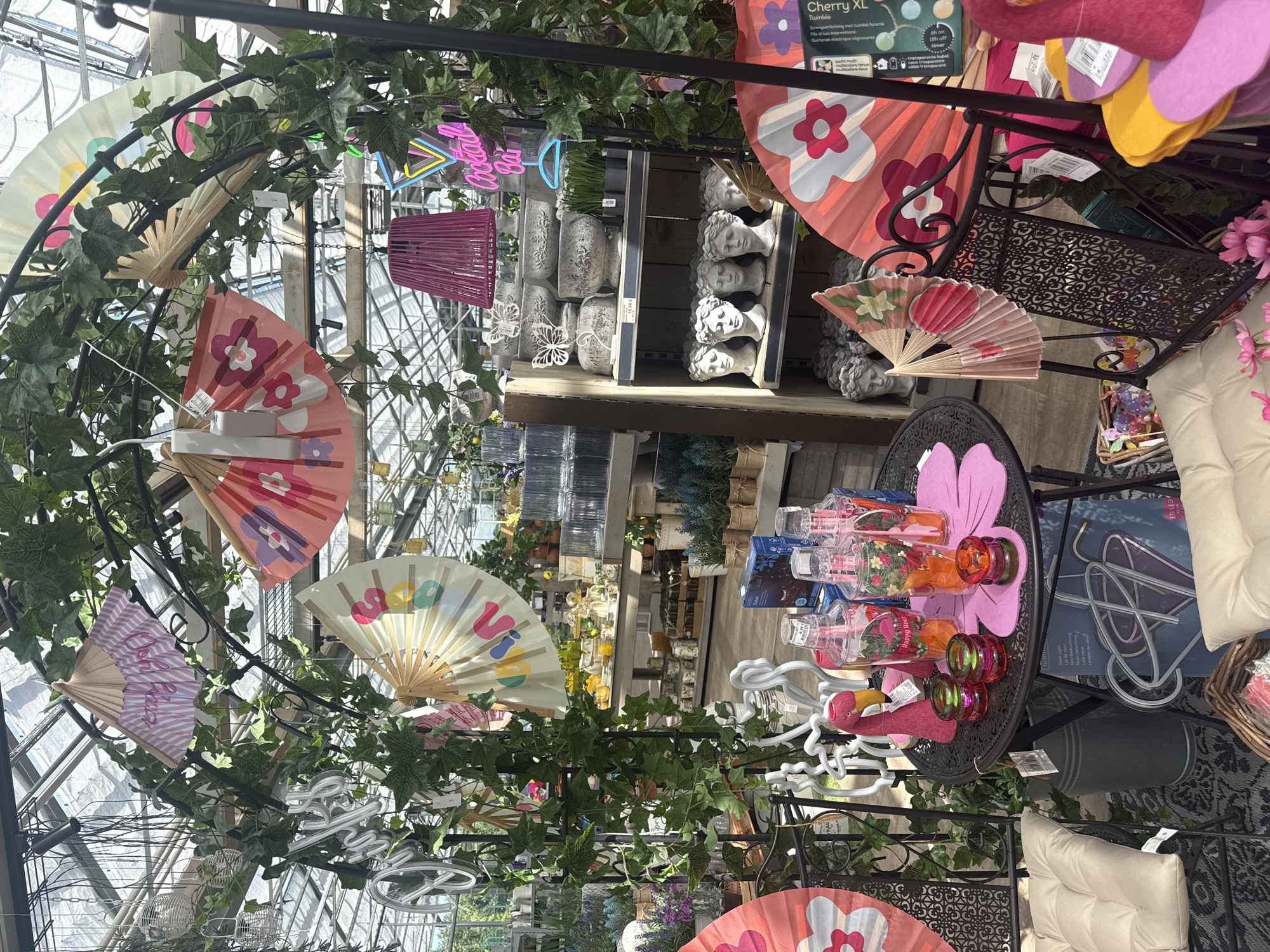 The image shows an outdoor market stall in Fritzlar, Germany, featuring a variety of decorative items, primarily fans and garden ornaments. The stall is adorned with ivy and other greenery, creating a somewhat whimsical atmosphere. In the foreground, a small round table displays several pink and orange plastic cups with straws, a pink flamingo figurine, and small glass candle holders. Behind the table, a dark metal chair is partially visible, decorated with a large pink fan featuring a floral pattern.
Further back, shelves are stocked with decorative pots and bust-like ornaments. A neon sign spelling "Cocktail Bar" is visible in the background, suggesting a festive or celebratory theme. Several other fans, in various sizes and colors, are hung from an arched structure covered in greenery. The overall impression is of a brightly lit, summery display of goods. The location is identified as Fritzlar, Germany, though the specific context of the market or the exact stall within the city is not provided. No people are visible in the image.