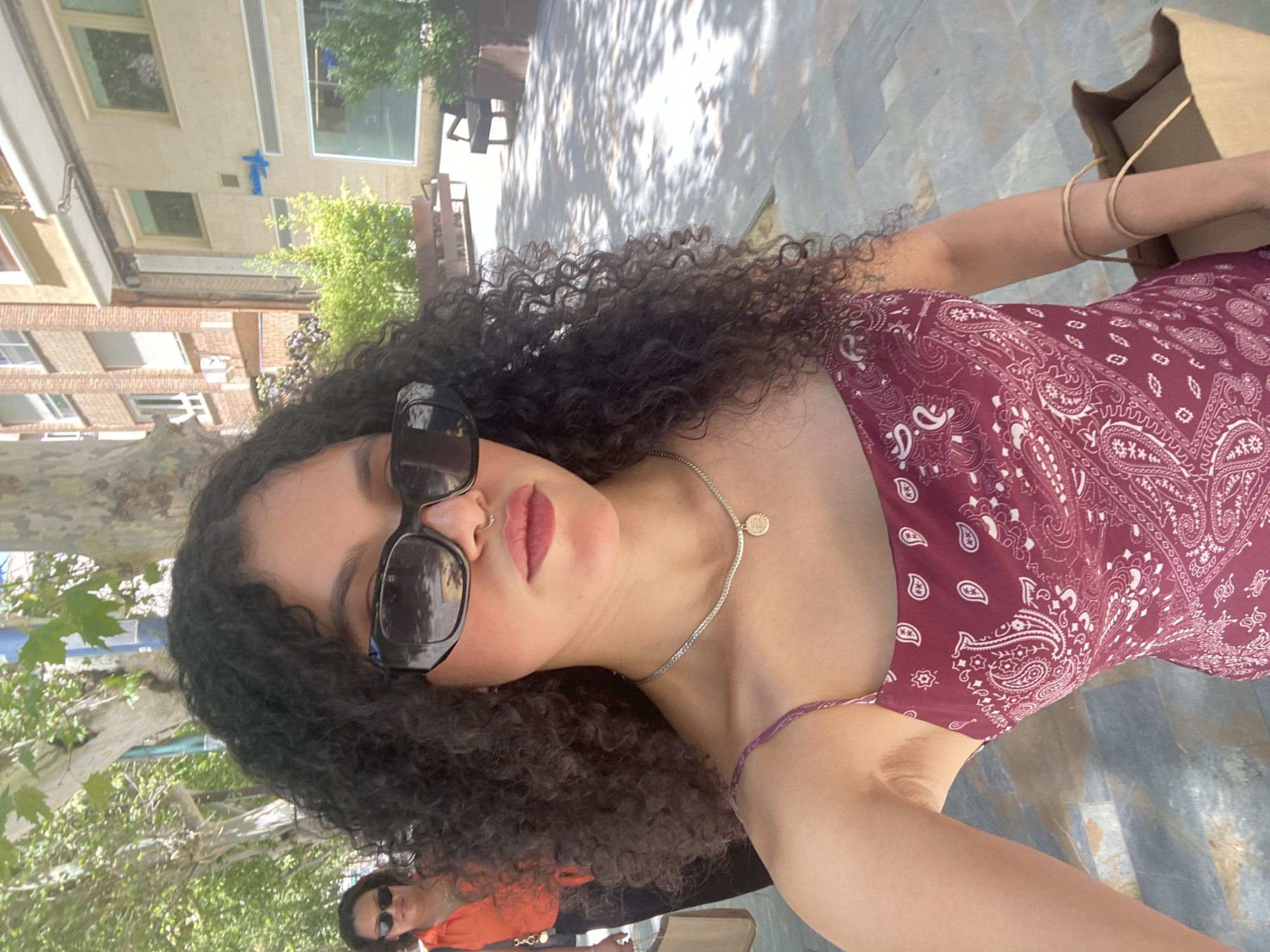This media file depicts a young woman with curly, dark hair, wearing sunglasses and a burgundy paisley-patterned dress. She is in a selfie-style shot, with her face close to the camera and her arm extending outwards, holding a brown paper bag. A second person, a woman wearing an orange top and sunglasses, is visible in the background, slightly out of focus.
The setting appears to be an outdoor urban environment. Hints of buildings, trees, and dappled sunlight on a paved surface suggest a daytime scene, likely in warm weather. The buildings in the background have a mix of brick and lighter colored facades, with windows visible. The paved ground is made of large, irregular stone slabs. The presence of a shopping bag implies the subjects might be engaged in or have just finished shopping.
The woman in the foreground has a relaxed and casual demeanor, with a slight smile. Her lips are painted a rosy pink, and she is wearing a delicate silver necklace with a small circular pendant. A thin, purple string bracelet is visible on her wrist. The lighting indicates it is daytime, with shadows suggesting the sun is present.
There is no visible text in the media file. The context of Murcia, Spain, suggests a warm, possibly Mediterranean climate.
