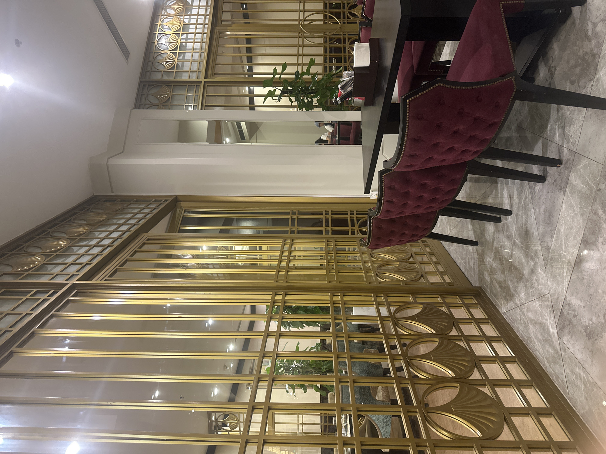 The image shows the interior of a dining area, possibly a restaurant or a private dining room. The focus is on a table setting with dark-colored tables and tufted, dark-red upholstered chairs. A plant serves as a centerpiece on the table. The flooring consists of gray marble tiles.
A notable feature is the extensive use of decorative gold-colored metal screens with geometric and circular patterns. These screens create divisions within the space and add a touch of elegance. Reflections in the screens suggest other parts of the dining area or additional seating.
The scene appears to be professionally designed with an emphasis on aesthetic detail and a sense of luxury. The lack of direct human presence, except for possibly a reflection of a person in the background, suggests the photo was taken to showcase the room's design. The image is sharp, highlighting the textures and patterns of the surfaces.