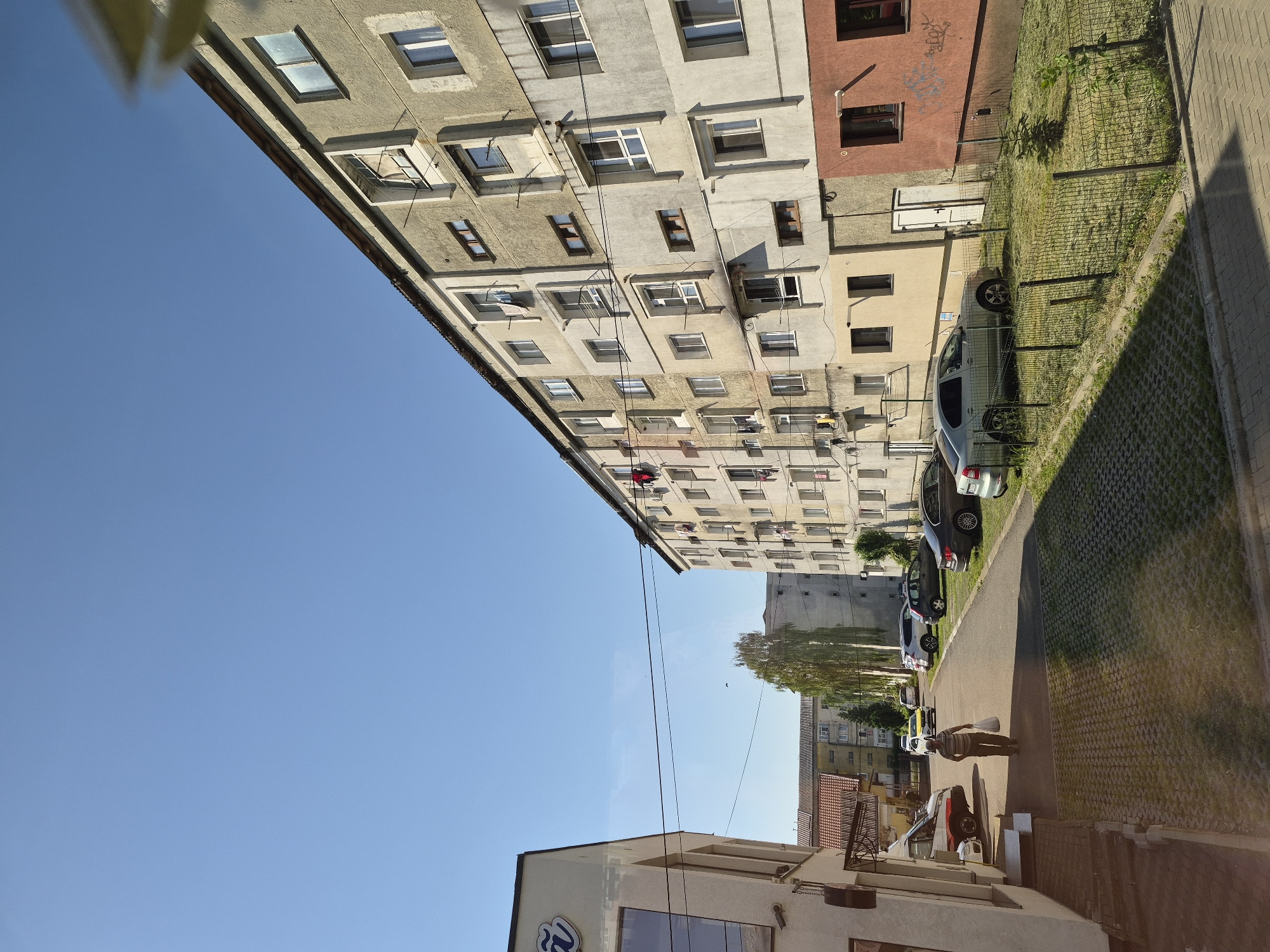The image depicts a residential street scene in Fălticeni, Romania, characterized by multi-story apartment buildings and parked cars under a clear blue sky. The foreground shows a paved area with interlocking pavers, transitioning to a walkway and a street.
On the left side of the image is a light-colored building with a prominent dark sign featuring a stylized blue 'Ñ' logo. A person is walking away from the camera on the walkway, carrying a white plastic bag. Further down the street, a white van with orange and red markings is parked.
The main focus of the image is a long, multi-story apartment building dominating the right side of the frame. This building, with its concrete facade, exhibits a somewhat weathered appearance. Several windows are visible, some with laundry hanging on clotheslines. Balconies and external brackets are also present.
Across the street from the apartment building, another similar apartment building is visible in the background, set further back. Adjacent to the main apartment building on the right, there is a section with a reddish-brown facade. A small, fenced-off grassy area with a young tree is situated in front of this section of the building.
Several cars are parked along the street, including a silver hatchback, a dark sedan, and a white SUV. The street itself is asphalt. The overall environment suggests a typical urban residential area.
