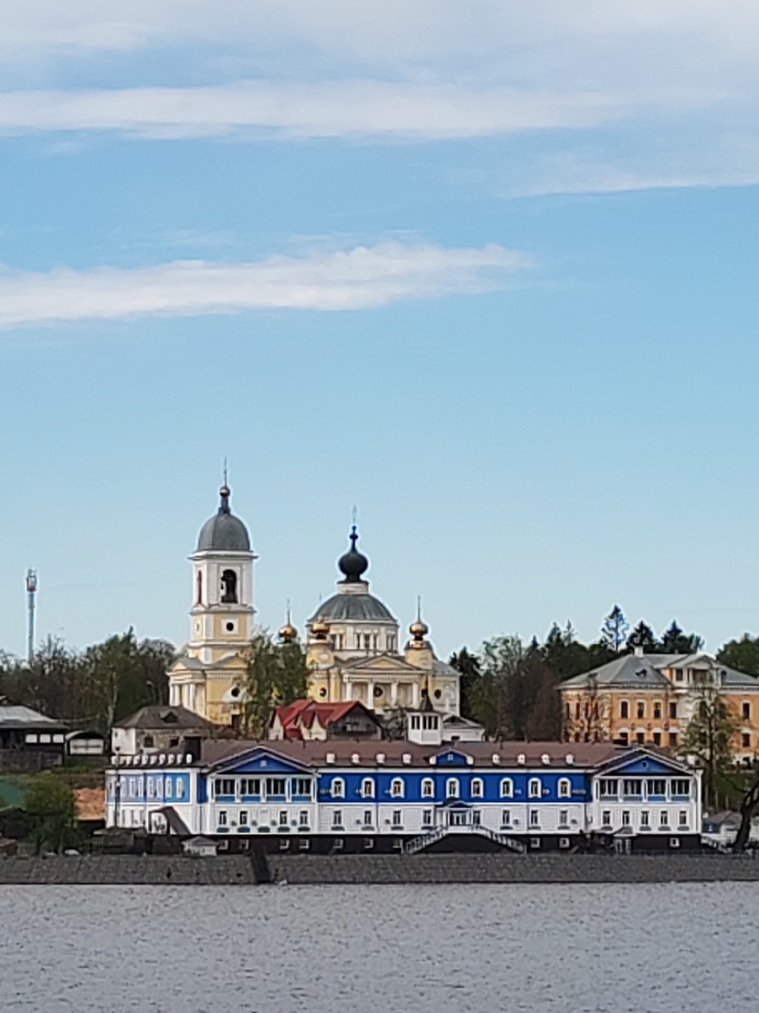 The image depicts a view of buildings and structures in Myshkin, Russia, likely taken from across the Volga River. A low-lying building with a blue and white facade is in the foreground, with stairs leading down to the river. Behind it, a yellow and white church complex with domes is visible, and further back, a large light yellow building. Green trees surround the structures. The sky is light blue with a few faint clouds. In the background, a telecom tower.