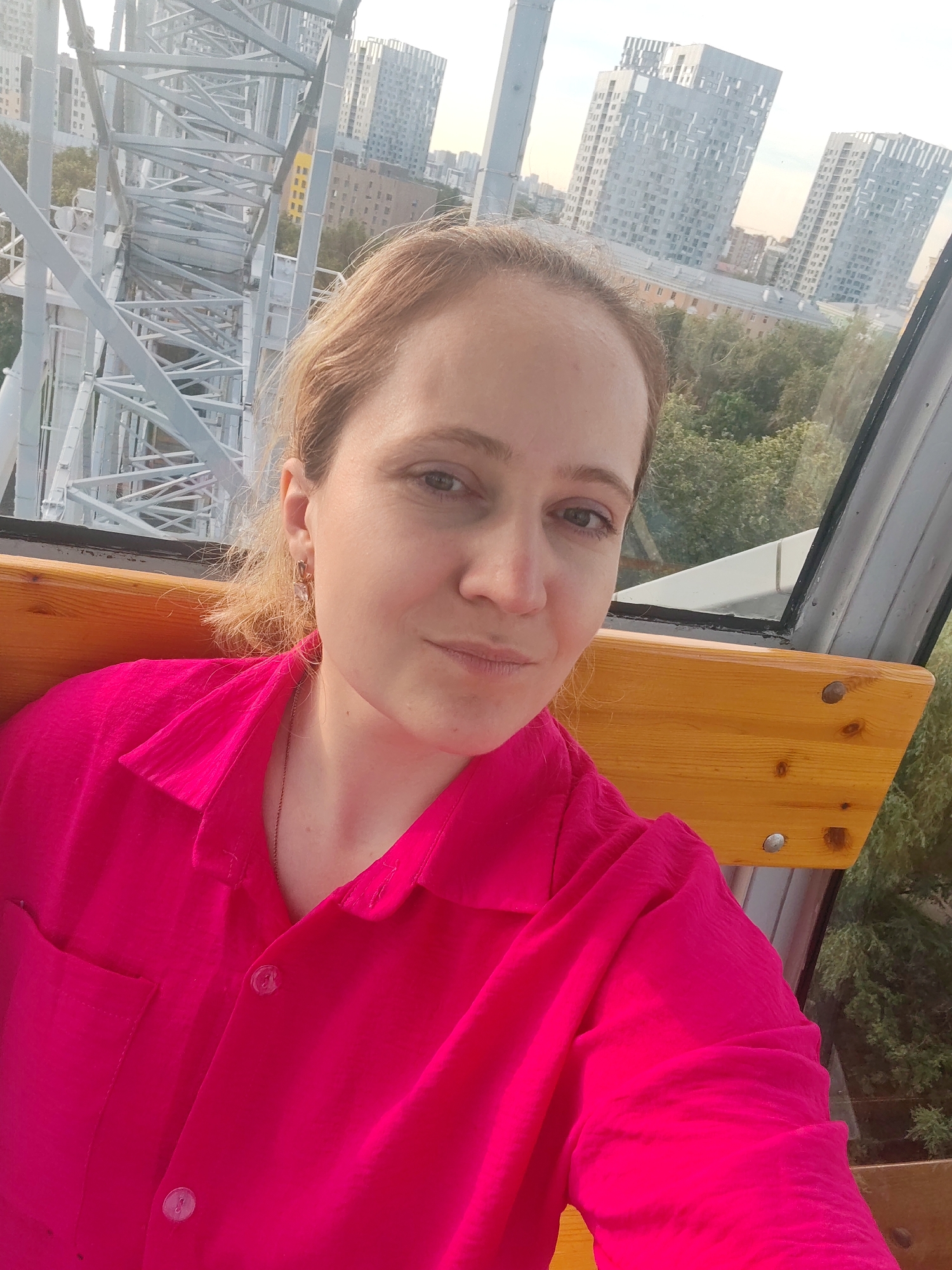 A woman with blonde hair and light skin is pictured in a selfie taken from inside a Ferris wheel gondola. She is wearing a bright pink button-down shirt and a thin necklace. She is looking towards the camera with a neutral expression. The background shows a portion of the Ferris wheel's structure on the left, and through the gondola window, a city skyline with modern buildings, trees, and a bright sky can be seen. The wooden bench of the gondola is visible in the foreground. This scene is likely within the "Permskiy Dvorik" (Perm Courtyard) amusement park or a similar location in the city of Perm, Russia, known for its Ferris wheel offering panoramic views of the Kama River and the cityscape.
