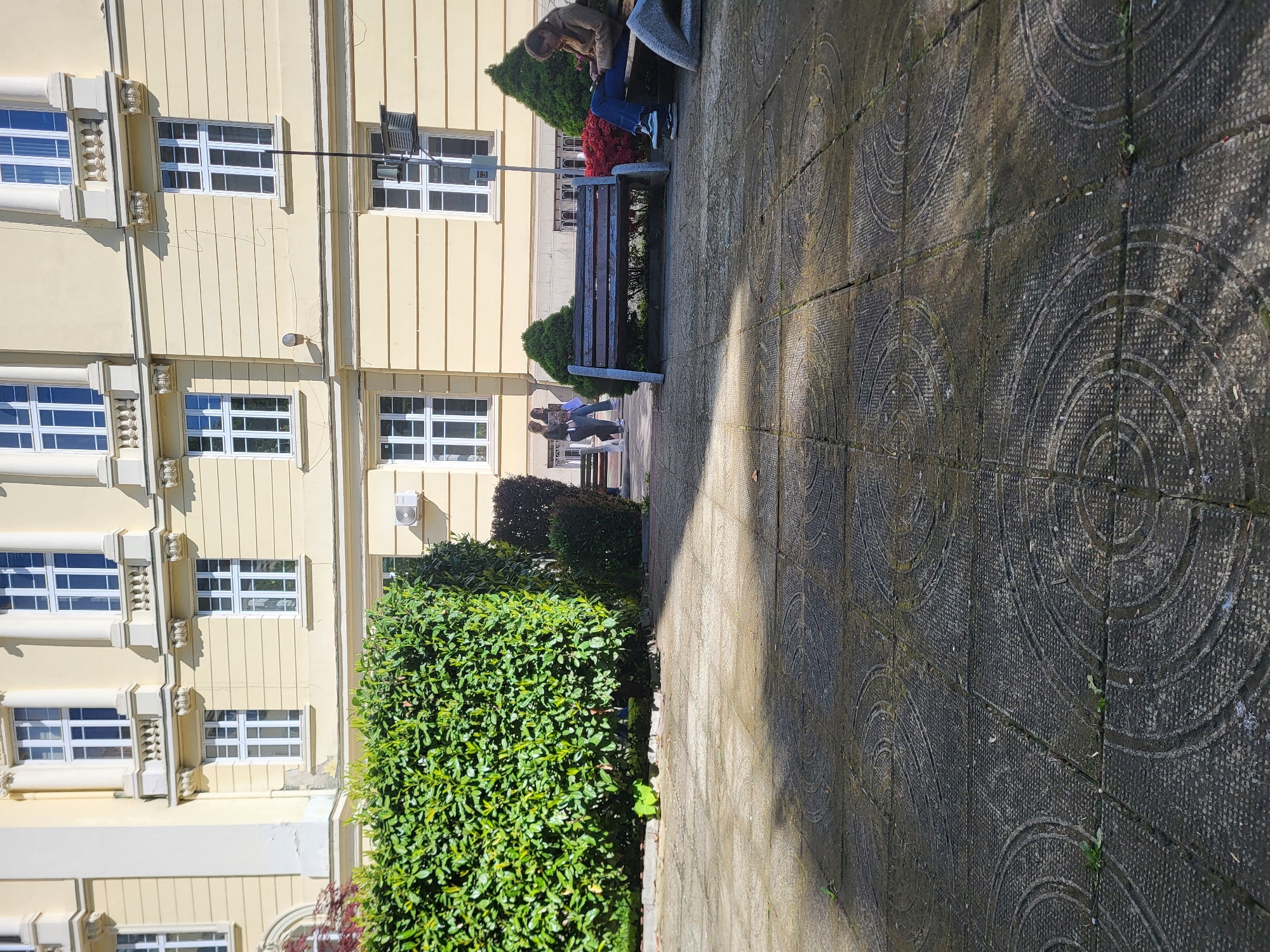 The image depicts a scene in Sofia, Bulgaria. The location appears to be a public space near a light yellow building with symmetrical white-framed windows and classical architectural details.
On the right side of the image, a patterned stone pathway stretches into the distance. A shadow bisects the pathway.
A bench is situated along the pathway. A person is sitting on one end of the bench. Further down the pathway, two people can be seen walking.
On the left side of the image, there are two hedges, varying in size. The building and the hedges line the pathway.