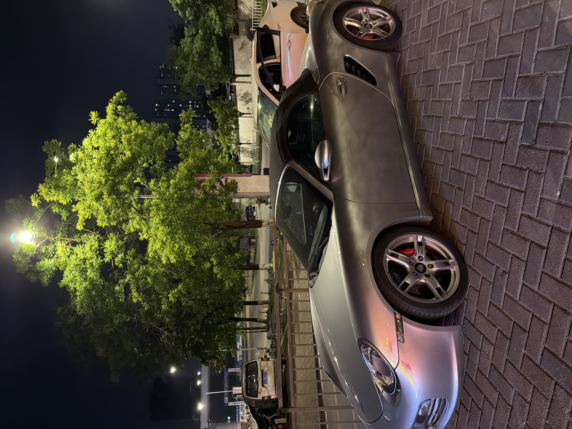 This nighttime image captures a street scene in Chongqing High-tech Zone, China. The primary subject is a silver Porsche Boxster convertible (likely a 987 generation) parked prominently on a dark, brick-patterned paved surface in the foreground. Its sleek metallic body reflects various light sources, including a bright street lamp on the left. Notable details on the car include multi-spoke wheels, through which red brake calipers with "PORSCHE" text are visible.
Behind the Porsche, a white SUV or van is partially parked, displaying a blue Chinese license plate with the characters "皖 A J7Q97" (皖 indicates Anhui province). The midground is dominated by large trees with vibrant green foliage, intensely illuminated by streetlights, particularly one on the far left of the frame.
The background shows a dark night sky and the silhouettes of urban high-rise buildings, some with scattered illuminated windows. There are no people visible, and no specific activity is taking place beyond the vehicles being parked. The scene is well-lit by artificial lights, creating a contrast between the illuminated trees and cars, and the dark sky and distant structures. The weather appears clear.
