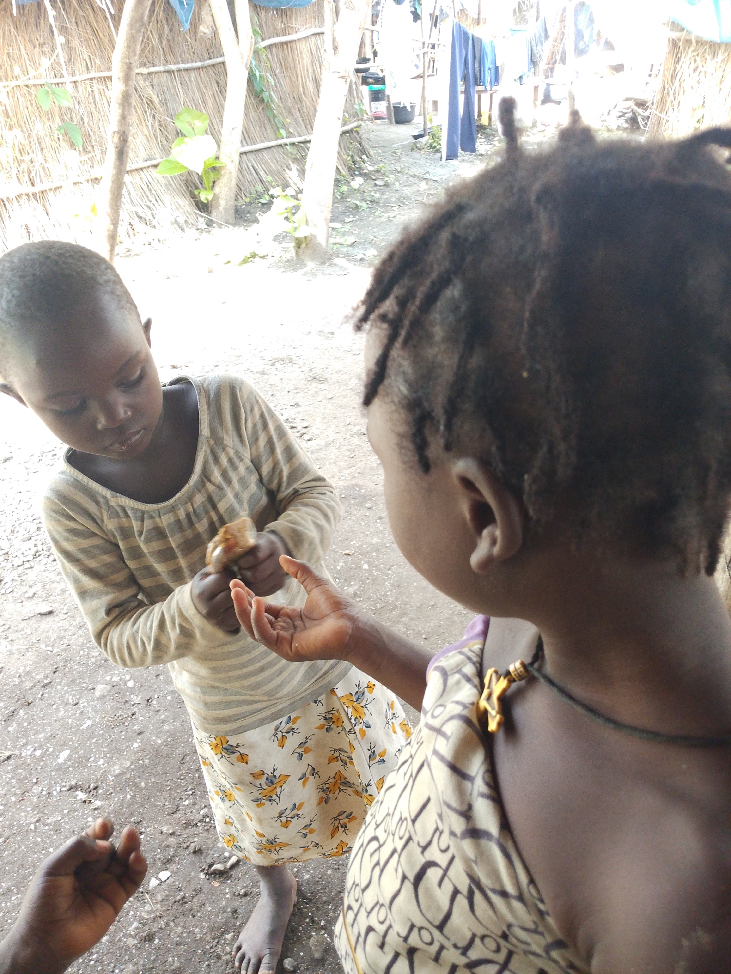 The image depicts an outdoor scene in what is identified as Otulu, Nigeria. Two young children are the central subjects. The child on the left, facing forward, wears a long-sleeved, striped shirt and a patterned skirt with yellow and blue floral designs. This child is holding a small, brown object, possibly food, in their hands, offering it to the other child. The second child, mostly visible from the back and side, has dark, braided hair and is wearing a patterned top with a necklace featuring a gold-colored pendant. The ground is covered in a mix of dirt and small pebbles. In the background, there is a structure made of vertically placed sticks and horizontal thatch material, suggesting a rustic dwelling or shelter. Some laundry hangs on a line in the distance, and vegetation is visible around the structure. The lighting suggests it is daytime, with bright sunlight casting some shadows.