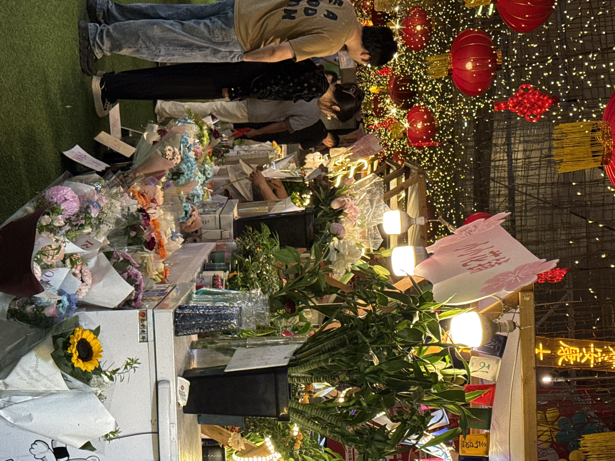 The image shows an outdoor display of flowers and other objects in what appears to be a commercial area. There are several people present, including a person in a beige shirt looking at the display. The background is decorated with lights and Chinese lanterns, suggesting a festival or celebration. The signage in Chinese and the presence of lanterns indicate that this is in China, specifically in Nan'an District based on context clues. There are various bouquets and arrangements of flowers displayed, as well as other objects and items.