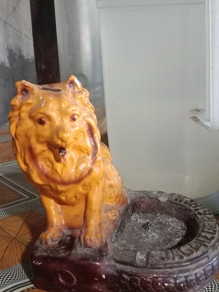 A weathered, yellow lion statue sits atop a round, dark-colored base. The lion has a coin slot on its head, suggesting it may be a piggy bank. The base appears to be an ashtray, filled with gray ash. The statue rests on a brown and tan patterned surface. A blurred white and gray background contrasts with the statue's prominent presence.