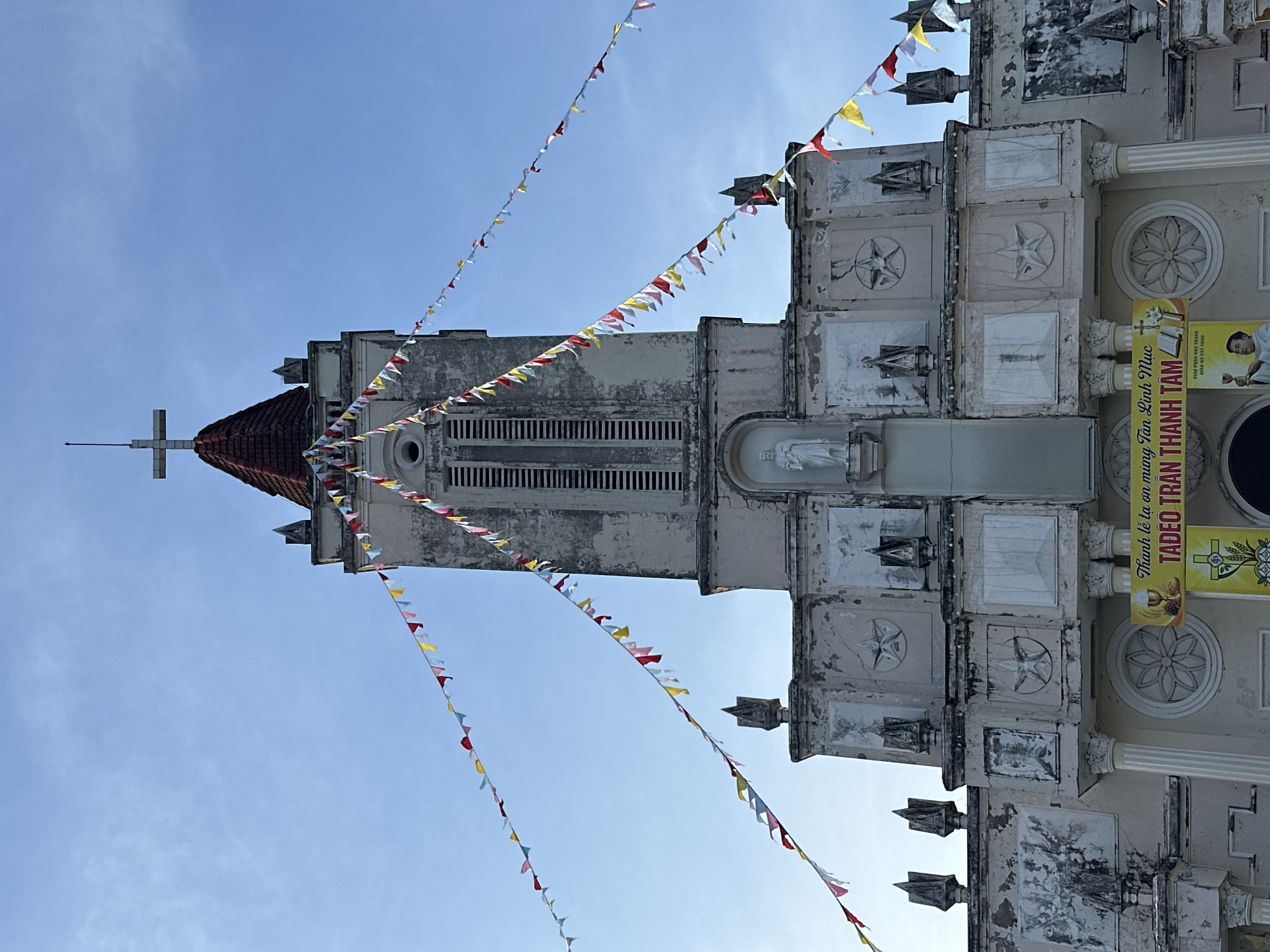 The image shows the front of a weathered church building under a clear blue sky. The church has a tall, square tower with a red, pitched roof topped by a cross. Colorful triangular pennant flags are strung across the front of the building. The facade of the church is adorned with decorative elements, including stars, religious iconography, and statues. There are pillars supporting the upper levels of the building. A yellow banner with Vietnamese text and religious imagery is displayed near the entrance. The overall condition of the building suggests it is an older structure. The location is identified as Diên Khánh, Vietnam, with the banner indicating a celebration or event related to a priest named "TADEO TRAN THANH TAM" and the local church.