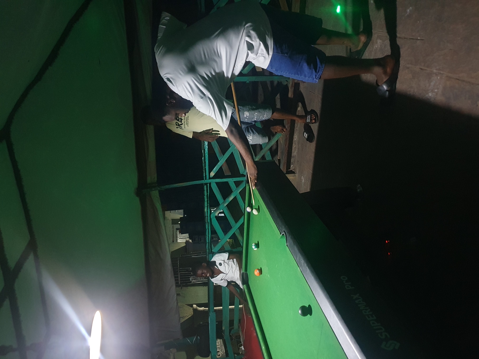 The image depicts an outdoor recreational area, possibly a bar or social gathering spot, located in Lokoja, Nigeria. The scene is illuminated by a bright overhead light and the ambient glow of a green-lit environment. In the foreground, a man is actively playing pool, holding a cue stick and aiming at a ball on the green felt of the pool table. He is dressed casually in a light-colored t-shirt and blue denim shorts. To his left, a young boy in a white polo shirt is seated, observing the game with a wide smile. Further in the background, another individual stands, partially obscured, wearing a yellow t-shirt. The environment features wooden structures and fencing, with a portion of the pool table bearing the inscription "SUPERMAX Pro". The ground appears to be paved or made of concrete. The overall impression is one of casual leisure and social interaction.
