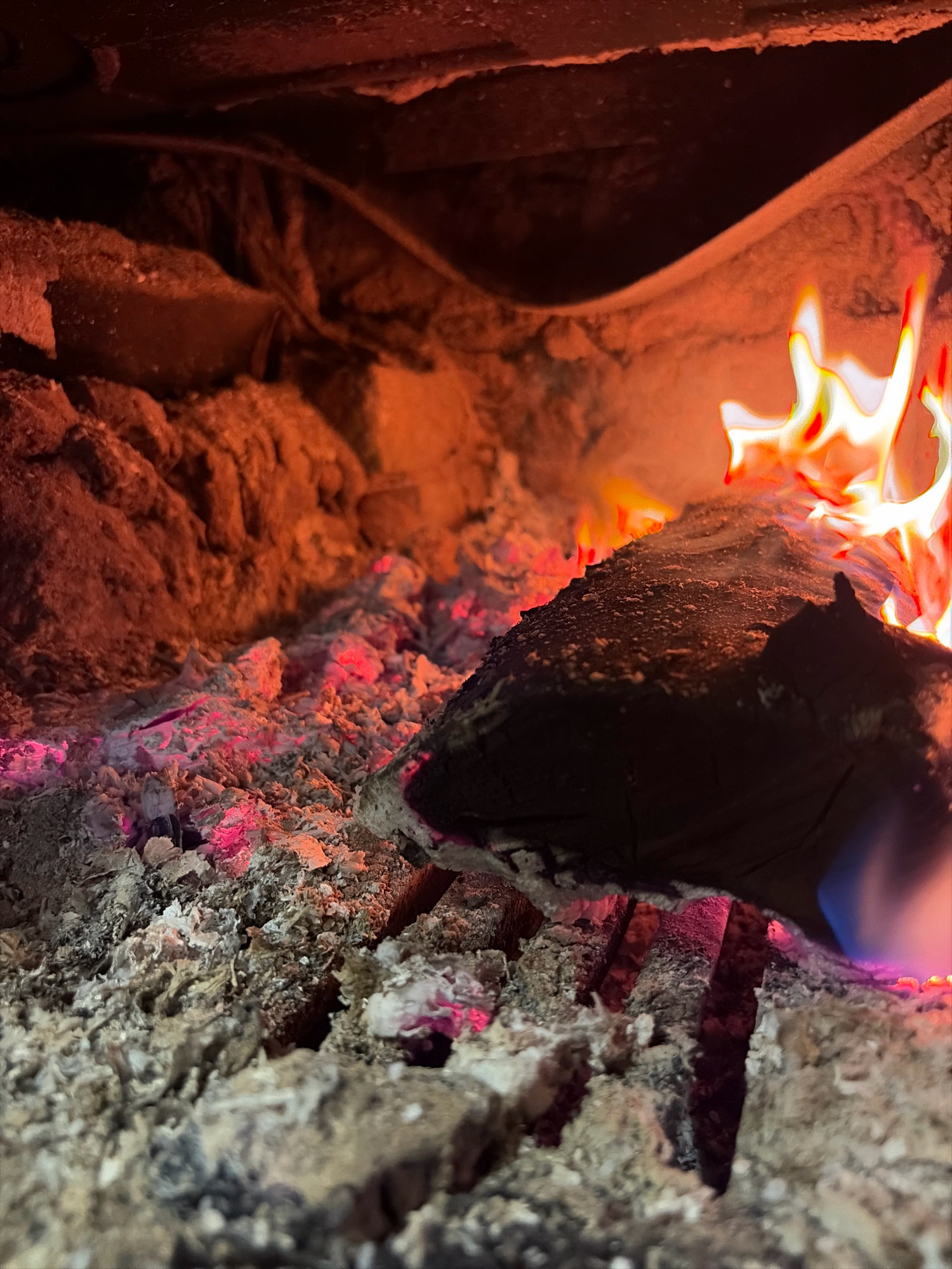 The image shows the interior of a fireplace with burning wood and glowing embers. The scene is dimly lit, with the flames providing the primary source of illumination. The environment appears to be a residential or outdoor setting, possibly a dacha, in Platforma 69-Y Km, Russia, given the context of the prompt. There are no people visible in the image. The focus is on the fire itself, with a large log of wood in the foreground, partially consumed by flames. The ash and embers are spread across the bottom of the fireplace, with some glowing red and pink. The background is dark and out of focus, with hints of the fireplace structure visible.