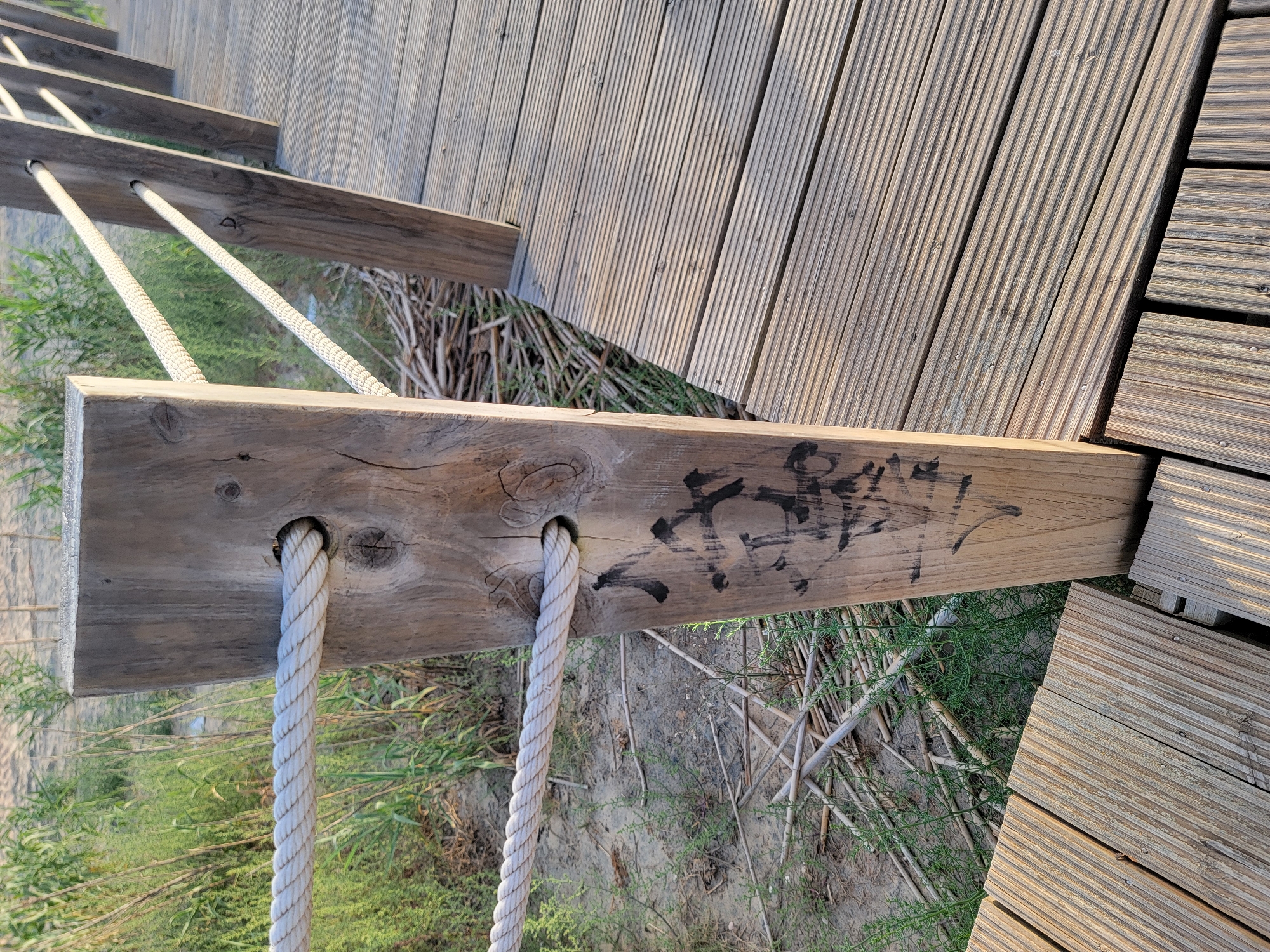 The image shows a wooden boardwalk post with rope railings and a wooden deck. The post is made of weathered wood and has two holes through which the ropes pass. There is black graffiti on the lower part of the post. The boardwalk appears to be located near a sandy area with vegetation. The scene suggests a coastal environment, possibly in Quarteira, Portugal, given the mention of the city.