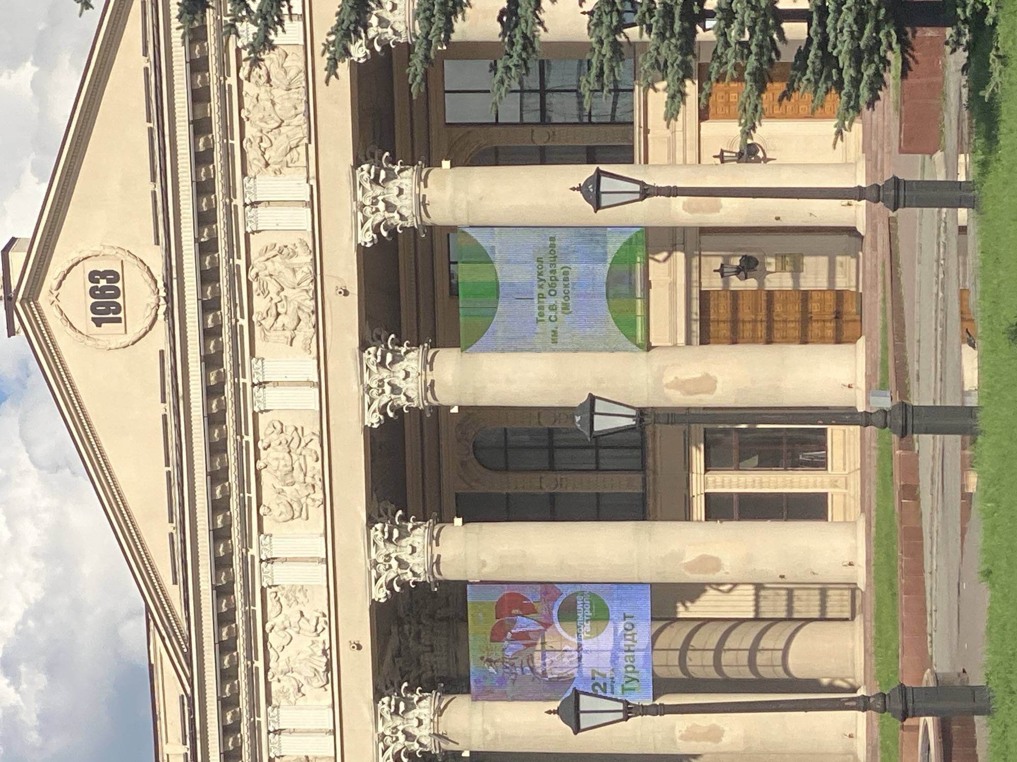 The image shows the facade of a building with classical architectural elements, including large columns and decorative friezes. A prominent feature is the year "1963" inscribed within a circular wreath above the entrance. Two banners are displayed on the columns; one on the left advertises "Turandot" with the number "27" prominently displayed, and the other on the right states "Театр кукол им. С.В. Образцова (Москва)" which translates to "Puppet Theatre named after S.V. Obraztsov (Moscow)". The building appears to be well-maintained, with lampposts positioned in front of the facade. The surrounding area includes a grassy lawn and a paved walkway. The sky is visible in the background with some clouds. This building is the Novokuznetsk Drama Theatre, located in Novokuznetsk, Russia.