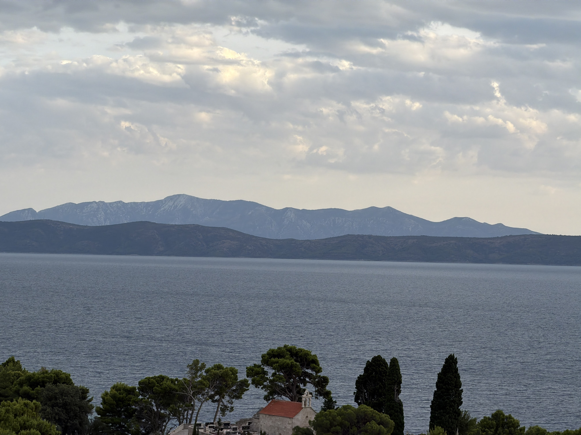 The image captures a panoramic view of the Adriatic Sea with a distant mountain range under a cloudy sky. In the foreground, a cluster of green trees and cypress trees frames a small, red-roofed building, possibly a church or chapel, situated in a graveyard with headstones visible. The scene is serene and appears to be located in a coastal area. Based on the visual elements, this could be a scene from Općina Podgora, Croatia, known for its coastal landscapes and historical structures.