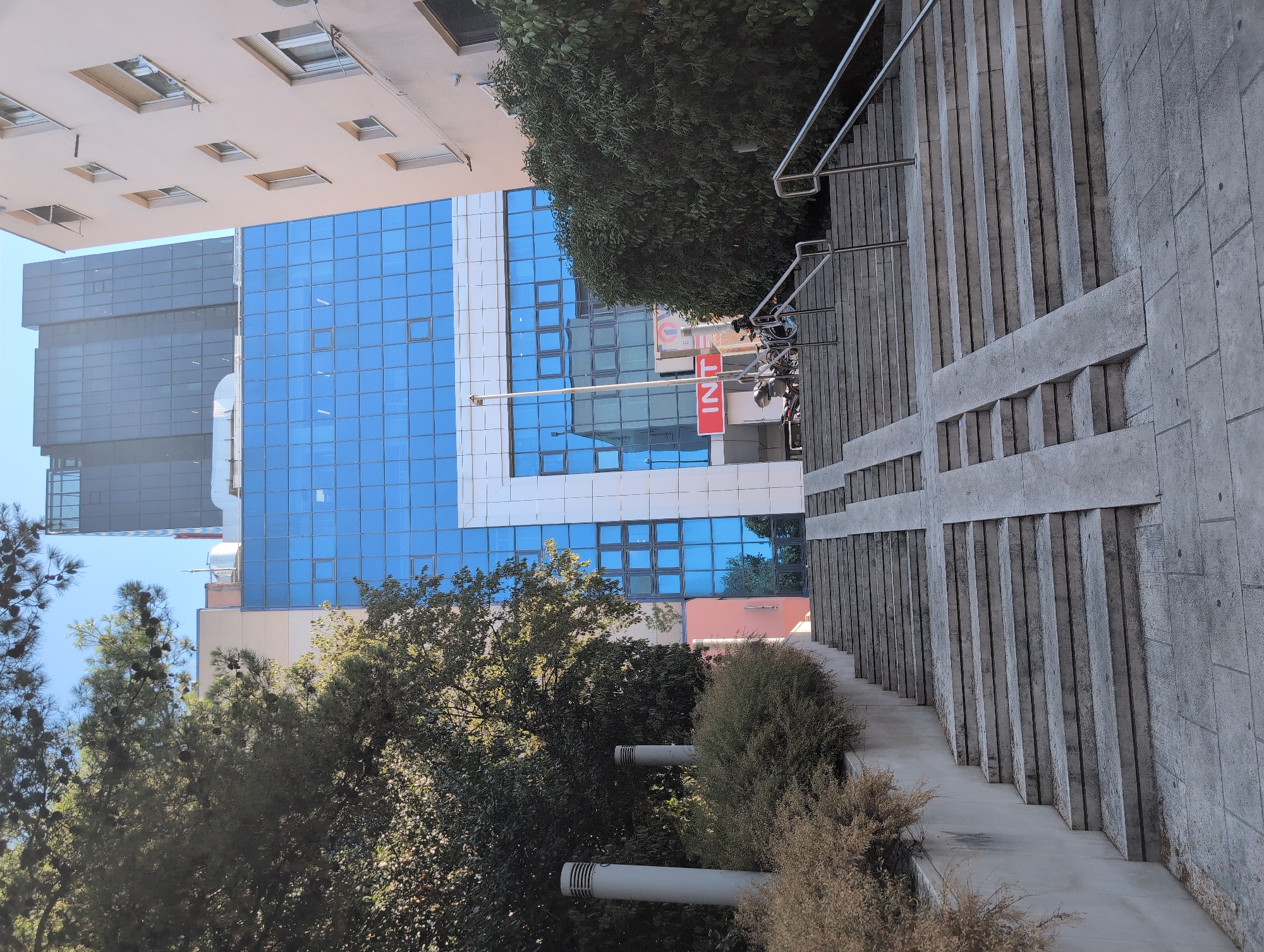 The image depicts an outdoor scene in front of a modern building with a blue glass facade and white accents. A wide concrete staircase with metal railings ascends towards the entrance. Integrated into the staircase are two ramps for accessibility. Lush green trees and bushes are visible on either side of the stairs and partially obscure the lower portion of the building. In the background, other buildings, including a darker, taller structure, are visible. A red sign with white lettering, partially obscured by greenery, is mounted on the blue facade. Two tall cylindrical light fixtures stand to the left of the stairs, near the bushes. Two bicycles and a person are visible near the top of the stairs, next to the entrance. The scene appears to be a public space, possibly a commercial or office building entrance. The location is identified as Grad Rijeka, Croatia.