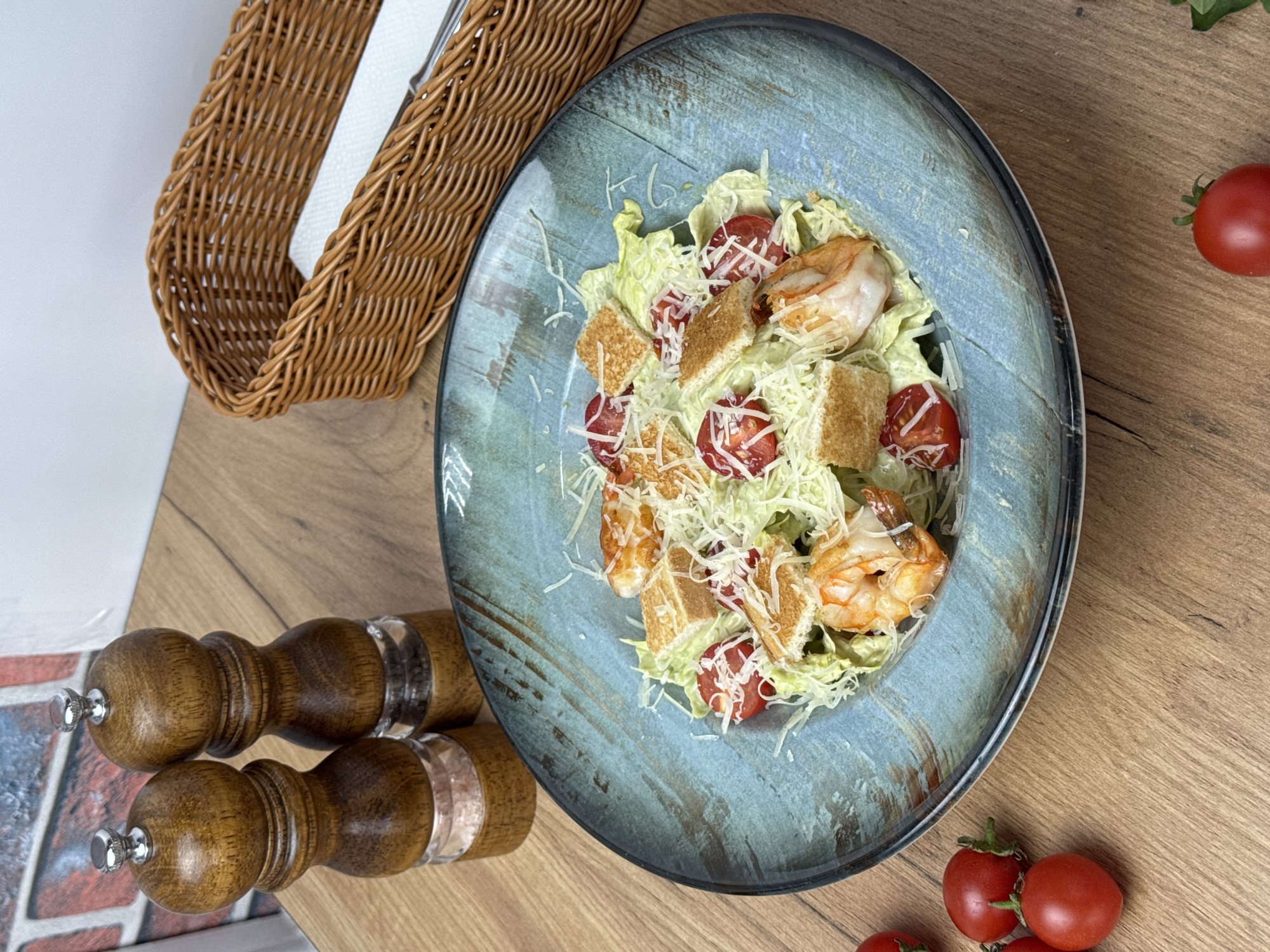The image depicts a plate of shrimp Caesar salad on a wooden table. The salad consists of lettuce, shrimp, croutons, tomatoes, and shredded cheese. Next to the plate, there are a pair of wooden pepper grinders and a wicker basket. In the background, a few loose tomatoes can be seen. The image was likely taken in a restaurant in Sochi, Russia.