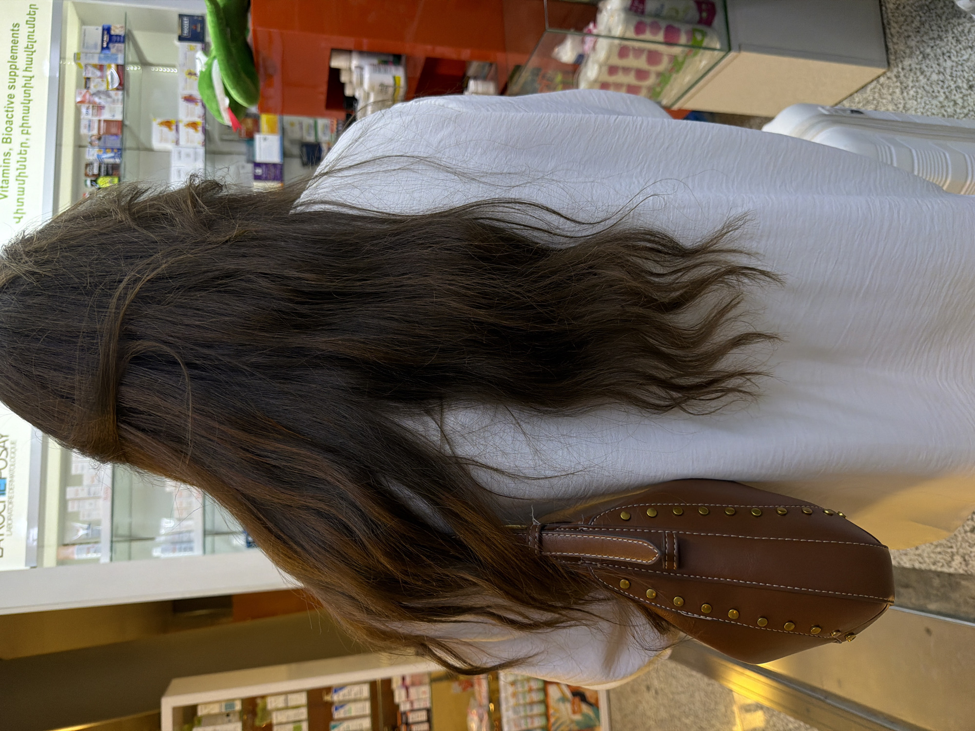 A woman with dark brown hair is standing inside what appears to be a pharmacy. She is wearing a white shirt or dress and has a brown handbag slung over her shoulder. Shelves stocked with various items, possibly medications or supplements, are visible in the background. Some text is visible on a product label, identifying it as "Vitamins, Bioactive supplements." The location appears to be in Yerevan, Armenia, based on the Armenian text present on the product labels in the background.