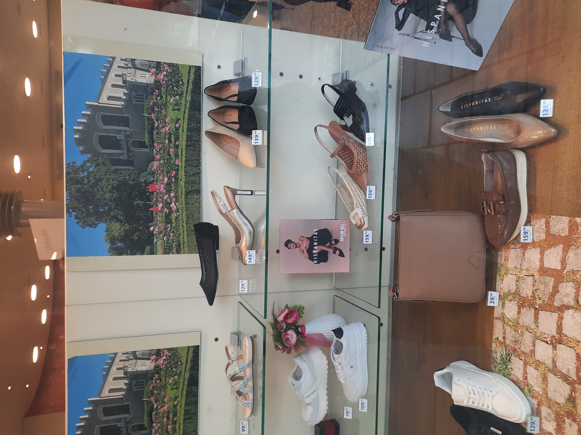This image shows a display window for a shoe store. The display features various shoes and a handbag. On the top shelf, there are black mesh flats, silver slingback heels, beige pointed-toe pumps, black pointed-toe pumps, and black slingback heels. The middle shelf has white sneakers, a vase with pink flowers, and a promotional poster for "HISPANITAS" featuring Taylor Hill. On the bottom shelf, there are white sneakers, a brown patent leather loafer, beige pointed-toe pumps, and black pointed-toe pumps. A light brown handbag is also visible on the bottom shelf. The store appears to be in Rosenheim, Germany, as suggested by the presence of the "palado" sign which is associated with a store in Rosenheim. The display window is well-lit, and the shoes are neatly arranged on glass shelves.