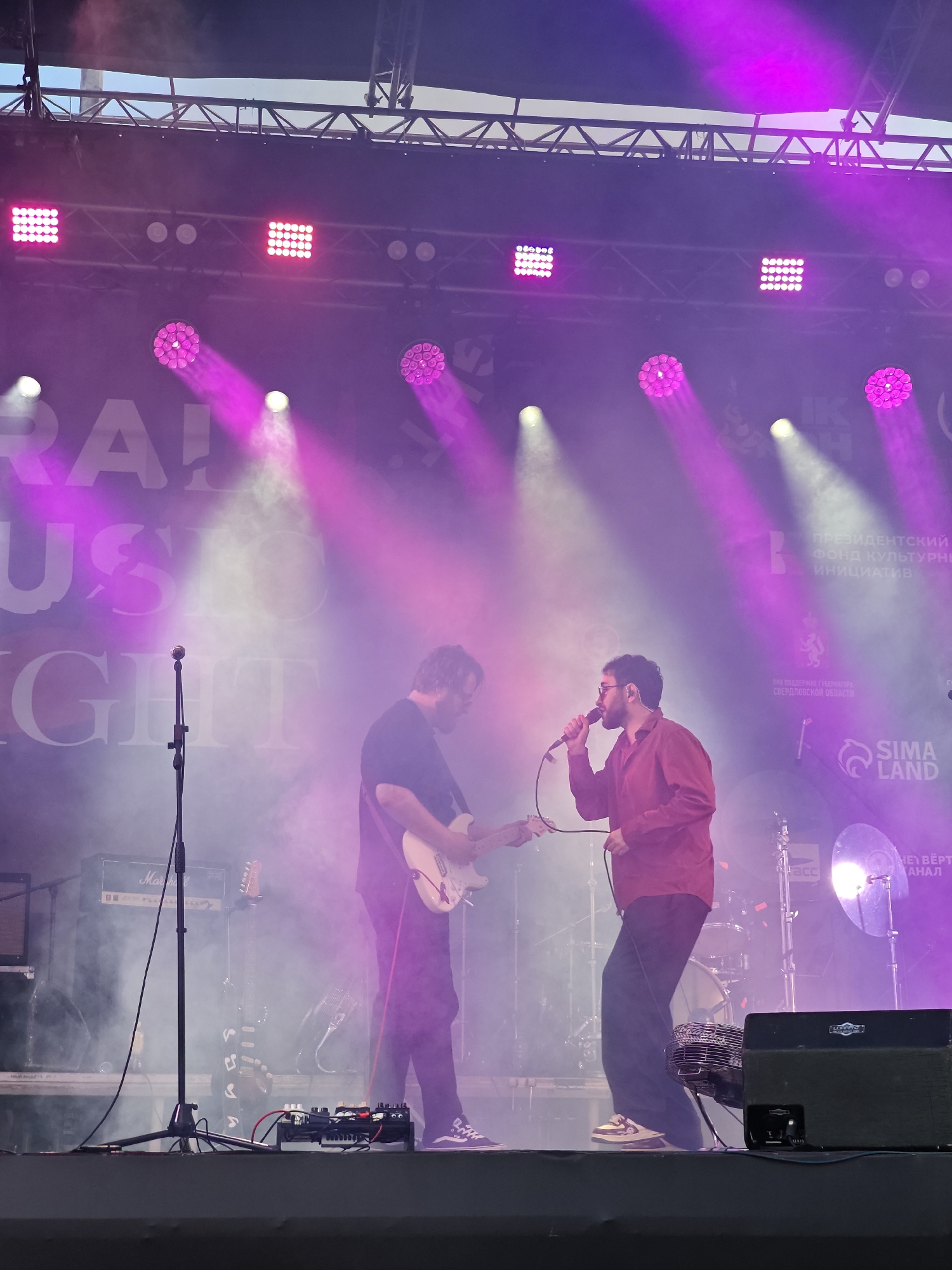The image depicts an outdoor music performance, likely at a festival, with two male musicians on stage. The musician on the left is playing a white electric guitar and wearing a dark t-shirt and dark pants. The musician on the right is singing into a microphone, wearing a reddish-brown button-down shirt and dark pants, and is also wearing glasses. They are illuminated by pink and white stage lights that create a smoky atmosphere. In the background, a large banner with white text, partially obscured by light and smoke, is visible. The text appears to include "RAI MUSIC NIGHT". To the right of the stage, logos and text indicate sponsorship or organizational support, with one recognizable logo reading "SIMA LAND" and another Cyrillic text indicating the "PRESIDENTIAL FUND FOR CULTURAL INITIATIVES" and support from the "GOVERNOR OF SVERDLOVSK REGION". The presence of the Sverdlovsk Region governor's support suggests the location is in Yekaterinburg, Russia. Various musical equipment, including amplifiers, microphones, and drum sets, are present on stage. A portable fan is also visible in front of the stage on the right. The overall scene captures a dynamic live music event.