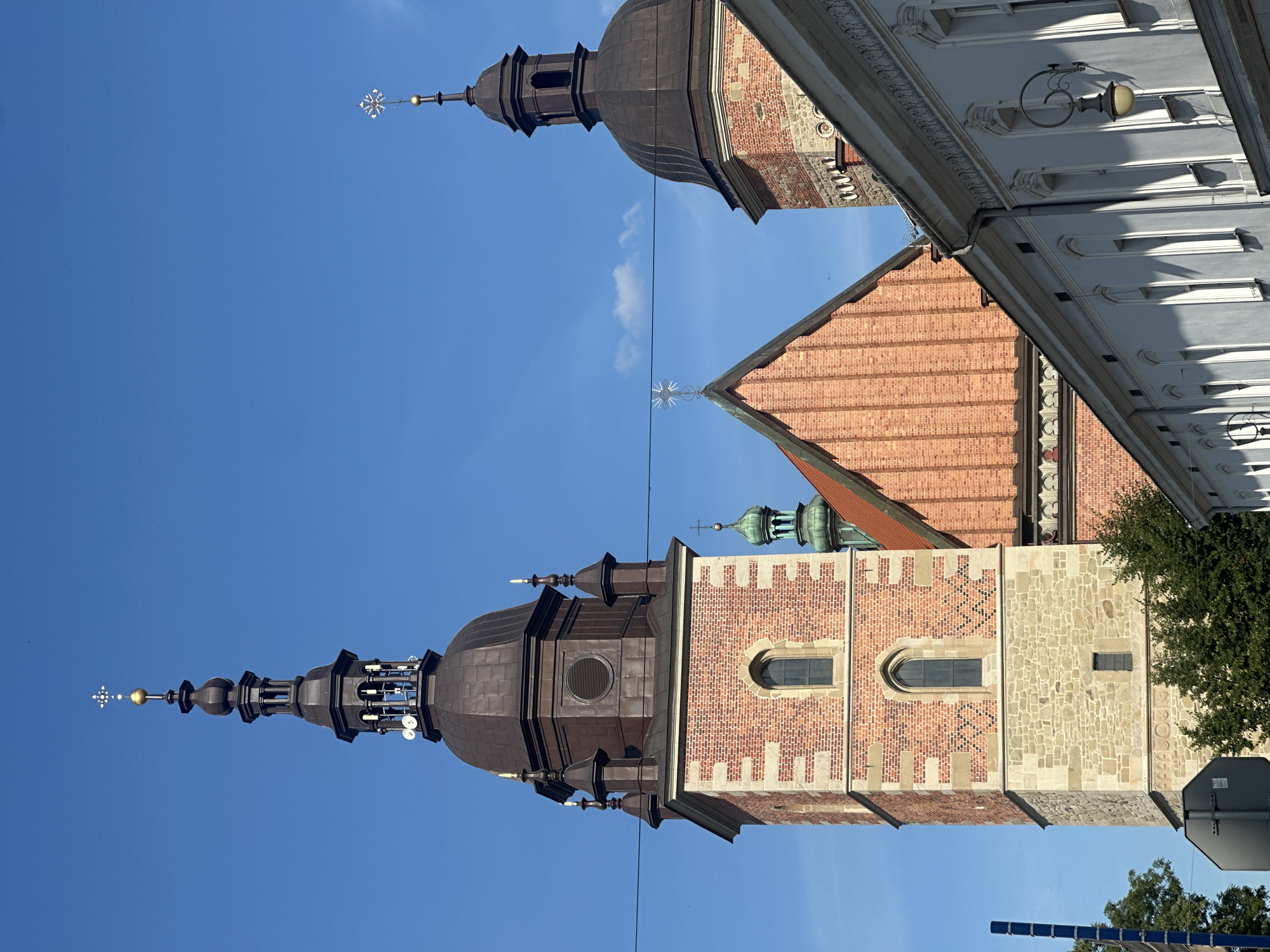 The image shows a view of the towers of St. Margaret's Basilica in Nowy Sącz, Poland, against a clear blue sky. The main tower, made of red brick and stone, is on the left. It features arched windows and a dark, domed belfry topped with a spire and a golden cross. To the right, another smaller tower, also with a dark dome and spire, is visible. A section of the basilica's red brick roof with decorative stonework is also in the frame. In the foreground, a white building with arched windows and decorative trim lines the street, casting shadows from the bright sunlight. The scene is framed by a power line running horizontally across the upper part of the image and a blue pole on the left edge. Trees are visible at the base of the main tower.