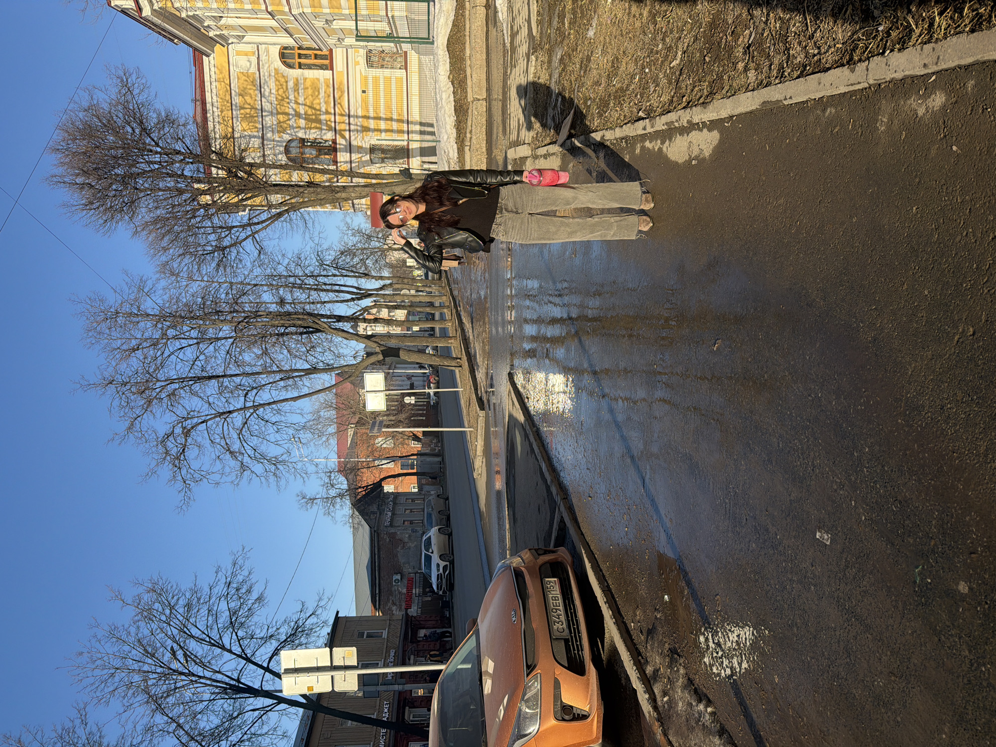 A street scene in Perm, Russia, features an unidentified woman with dark hair wearing sunglasses, a black leather jacket, and jeans. She is standing on the sidewalk next to an orange car parked on the right side of the street. She is holding a pink cup in her left hand, and she is raising her right hand in a gesture. The street is wet and reflects the sky. Bare trees line the street, and buildings of varied architecture are on either side. A traffic sign is visible. The license plate on the car is C349EB152.