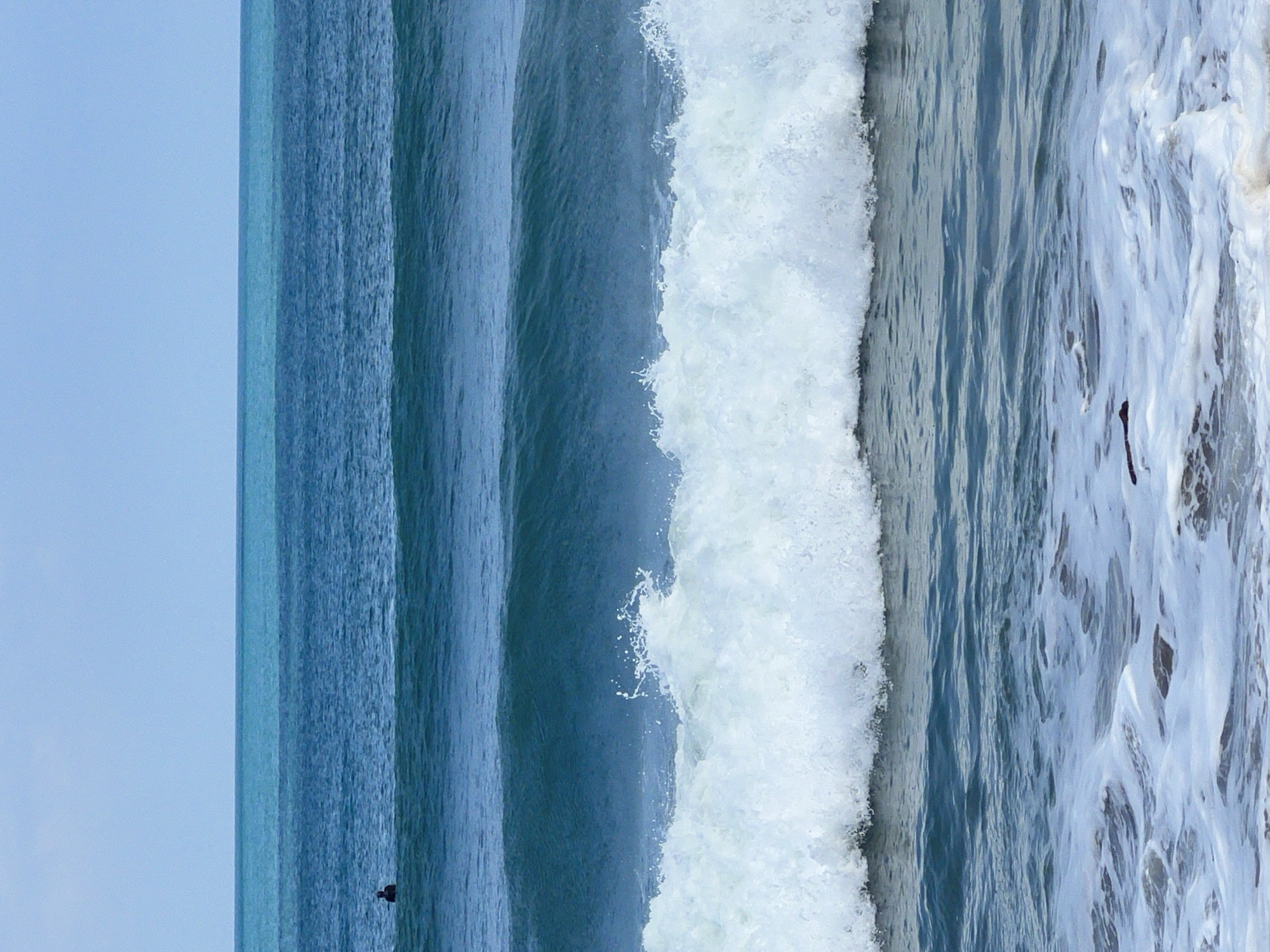 A wide shot captures a serene ocean scene with distinct layers of blue water under a pale sky. The horizon is sharply defined, separating the sky from the ocean. Close to the horizon, a strip of lighter blue water suggests shallow depth or a different water composition. Below this, a wider band of deep blue ocean stretches, with a lone dark figure in the distance, possibly a surfer, positioned on the surface. Further down, a prominent wave is captured mid-break, its crest frothing into white spray. The foreground features the receding white foam of smaller waves and the dark, wet sand of the shore, with a piece of dark debris visible in the foam. The image portrays a tranquil moment on the coast of Kuta, Indonesia, emphasizing the vastness and dynamic nature of the sea.
