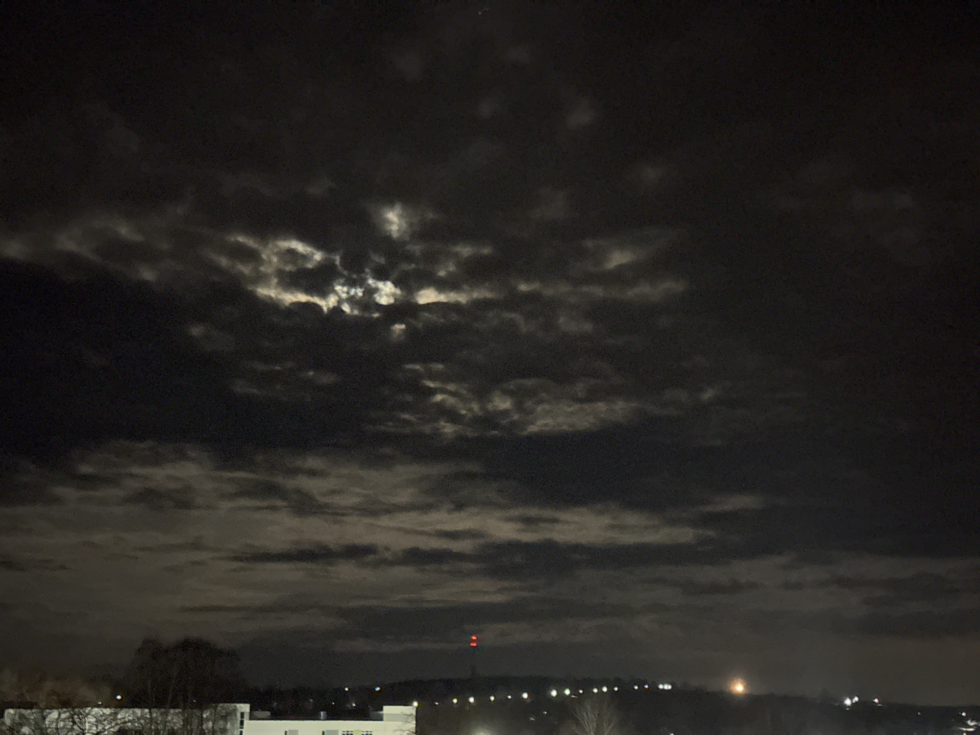 The image depicts a night scene in what is likely Chaikovsky, Russia, given the prompt's location. The sky is predominantly dark, filled with thick cloud cover, some areas of which are illuminated, likely by moonlight. In the foreground, the roofs of buildings are visible in silhouette. A line of distant lights suggests a road or street further away. In the middle ground, the faint outline of trees is discernible. A distant structure with a red light on top is also visible on the horizon. No people or clear actions are apparent in the image. The overall environment is dark, urban, and cloudy.
