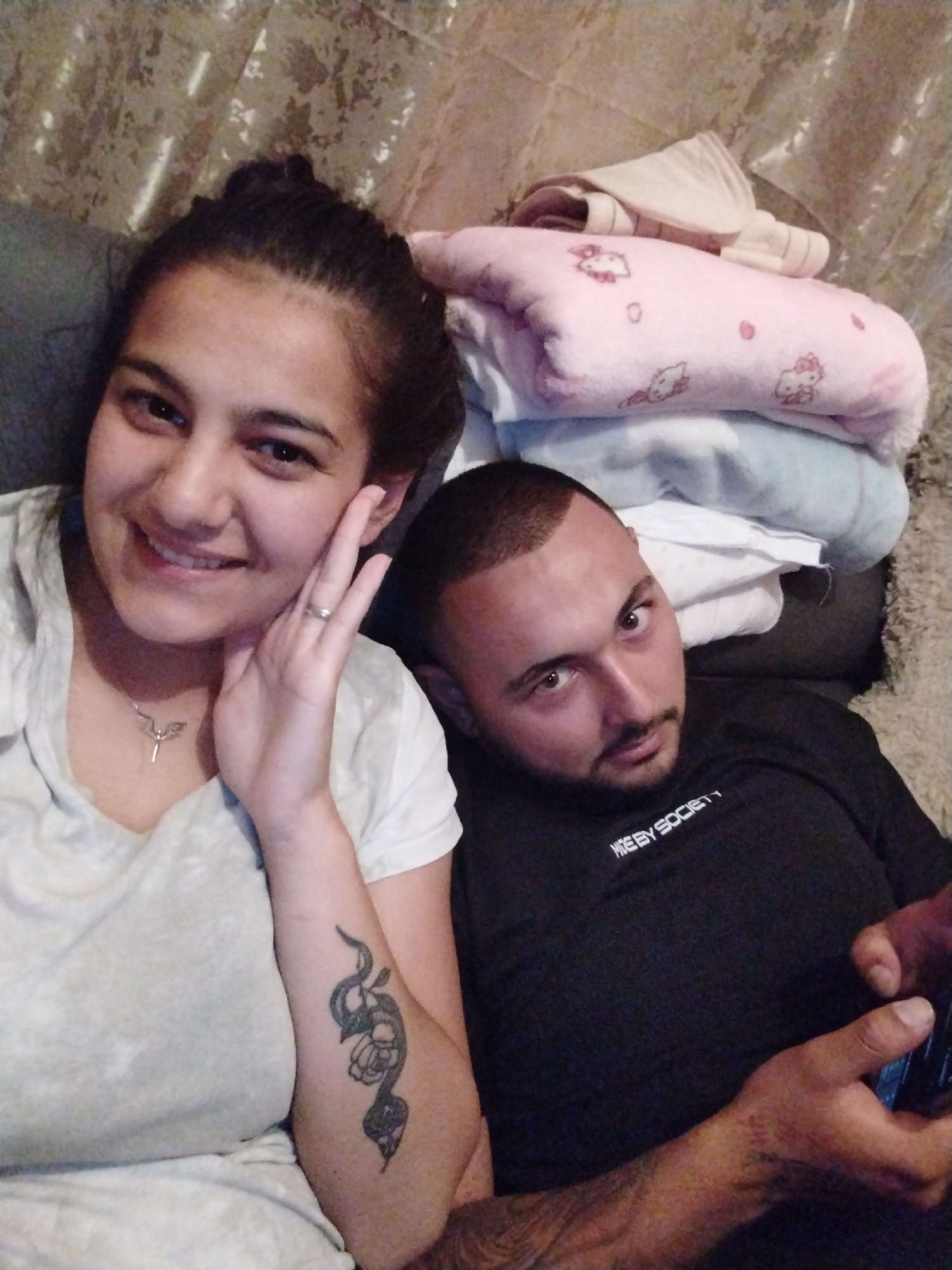 The image depicts two individuals indoors. A woman with dark hair pulled back, wearing a light-colored shirt and a necklace, is seated and smiling. She has a snake tattoo on her left arm. A man with a close-cut haircut, wearing a black shirt with white lettering that reads "Hype By Society", is lying next to her. He is looking at the camera while holding a device in his hands. Behind them, there are several folded blankets, including one with a Hello Kitty pattern. The background appears to be a wall with a textured covering. The location and city are unknown.