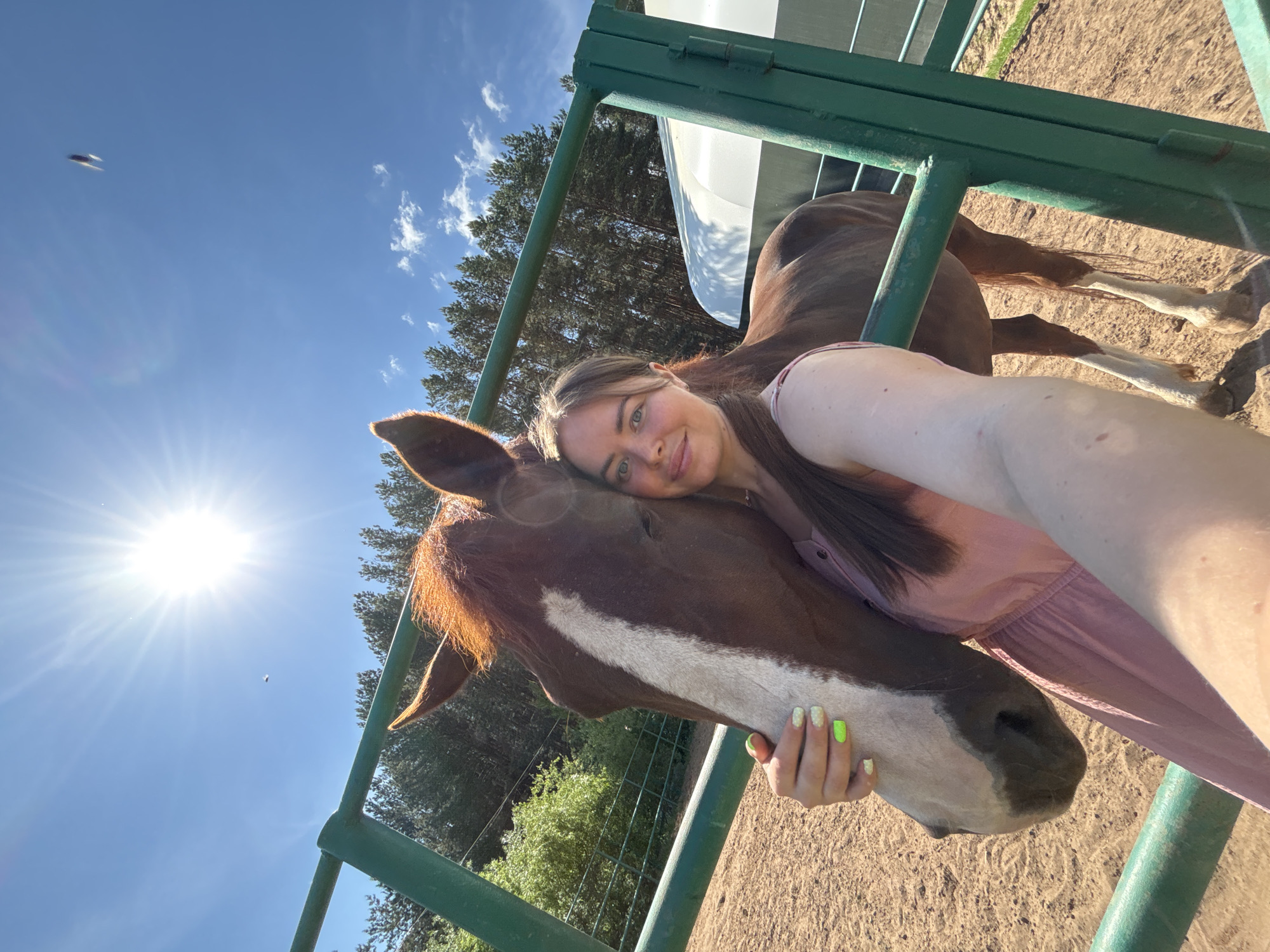 The image is a selfie taken outdoors on a sunny day. A fair-skinned woman with long brown hair, wearing a pink top, is embracing a brown and white horse. The horse has a white stripe down its face. They are positioned behind a green metal fence, with a sandy ground visible below. Trees are visible in the background. A white structure can be seen behind the fence. There is an object in the sky, above the horse's ear, that is possibly a bird. The city of Русский Вожой, Russia is not discernable from the context of this image.