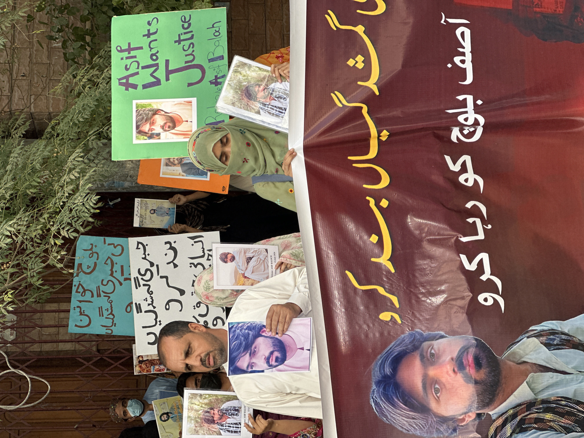 The image shows a protest in Quetta City Tehsil, Pakistan. A group of people are holding up signs and photographs. The signs are written in both English and Urdu. One sign in green reads "Asif Wants Justice". Another sign in blue reads "بلوچ خواتین کی جبری گمشدگیاں بند کرو" (Stop the enforced disappearances of Baloch women). A large banner in the foreground reads "گمشدگیاں بند کرو" (Stop disappearances) and "آصف بلوچ کو رہا کرو" (Release Asif Baloch). People in the image appear to be of Baloch ethnicity. The environment appears to be an urban setting, with a building in the background. The protest is likely related to human rights abuses and enforced disappearances in the Balochistan region.