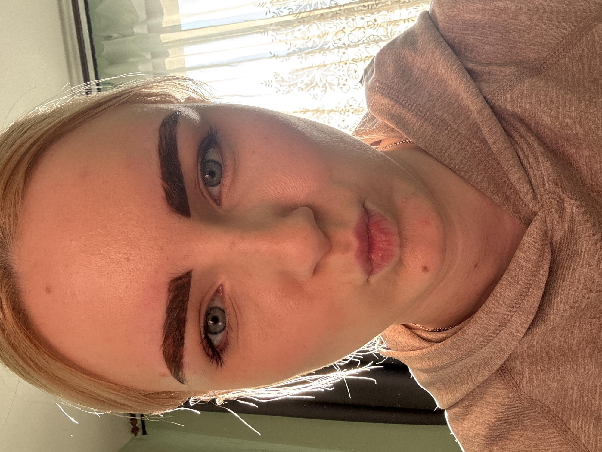 A close-up selfie of a young Caucasian woman with blonde hair pulled back shows her face from the eyebrows down to her shoulders. She has light blue eyes and dark, thick eyebrows. Her lips are pursed, and she is wearing a light brown hooded sweatshirt. The lighting suggests the photo was taken indoors during the daytime, possibly near a window, with sunlight casting shadows and highlighting her face. The background is blurred, showing hints of a curtain and a wall. The location is Unknown, Russia.
