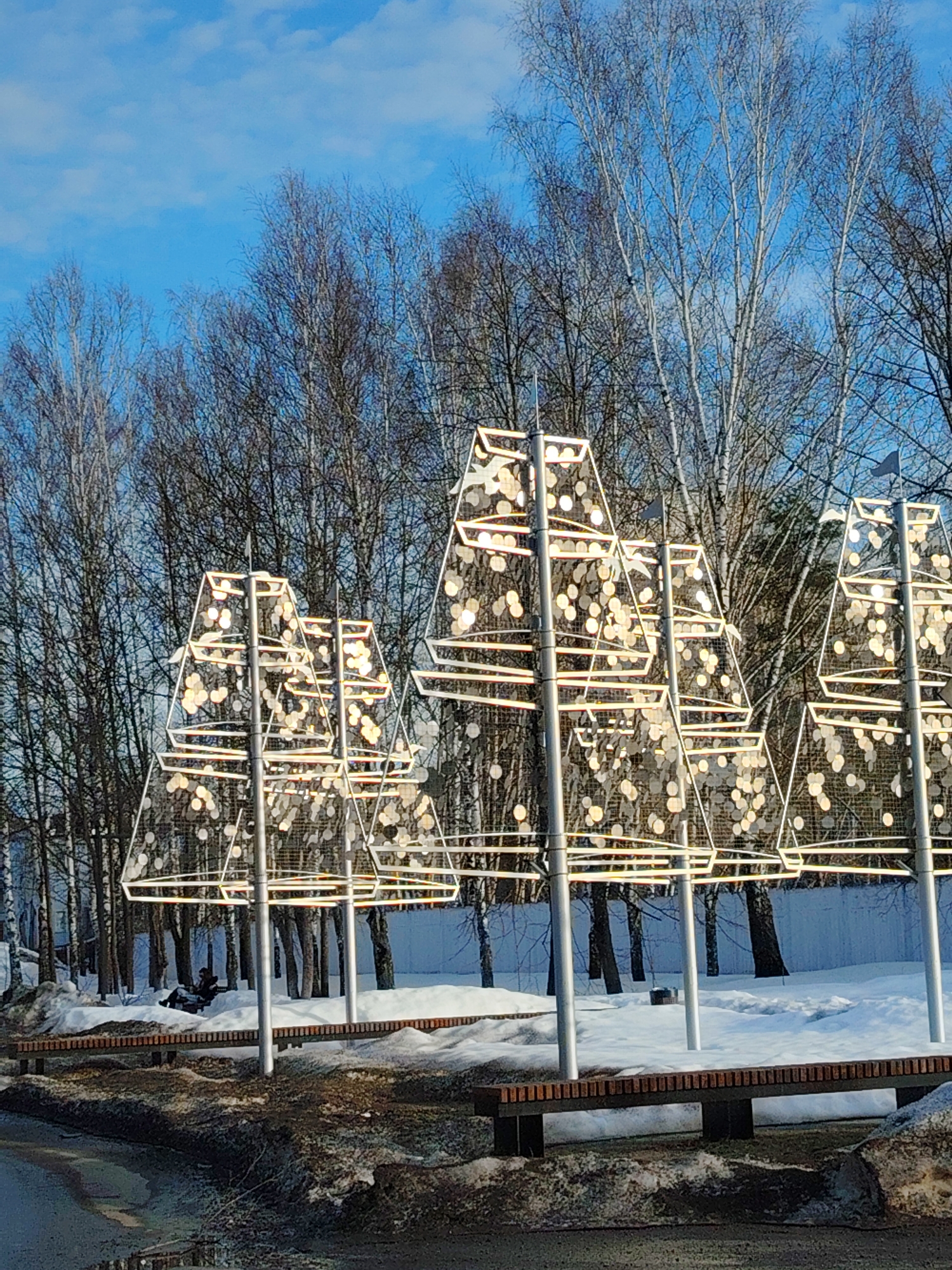 The image depicts an outdoor park or public area in Chaikovsky, Russia, under a clear blue sky with scattered white clouds. The scene is characterized by melting snow and ice on the ground, indicating a winter or early spring season.
In the mid-ground, a series of five prominent, stylized conical or pyramidal decorative structures are visible. These structures consist of light-colored metal frameworks with mesh infill, densely adorned with numerous circular, golden-hued reflective elements that brightly catch the sunlight. Each structure is supported by a slender pole, and several are topped with small, dark pennant-shaped flags.
Behind these decorative elements, a dense line of bare deciduous trees, including several identifiable birches with light-colored trunks, forms a natural backdrop. The foreground features a dark asphalt path or road, partially covered with melted snow, dirt, and puddles.
To the left of the image, in the mid-ground, a solitary individual is seated on a dark-framed wooden slatted bench. This person is dressed in dark winter clothing. Additional similar benches are partially visible along the lower right side of the frame, some partially obscured by snowdrifts. The ground around the benches and decorative structures is covered with patches of snow of varying depths and cleanliness.