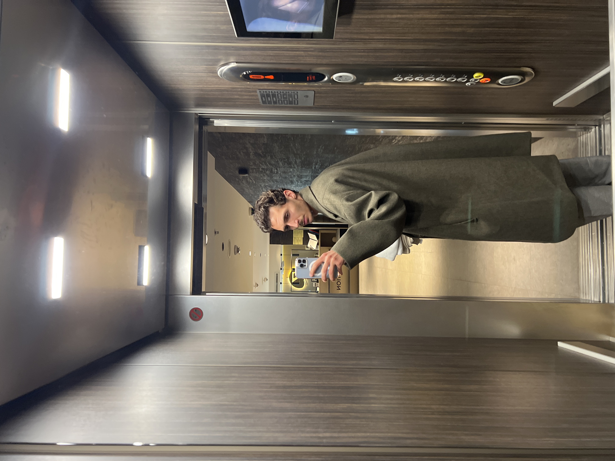 A man in a gray coat is taking a selfie in an elevator. The elevator has wood-paneled walls, bright rectangular lights on the left wall, and a panel with floor numbers and buttons above the elevator door. The elevator is stopped at a floor with a view into a brightly lit space with clothing racks and a sign that appears to say "HOM". The man is holding a light blue iPhone and has short brown hair. The location may be a clothing store in Budva, Montenegro.