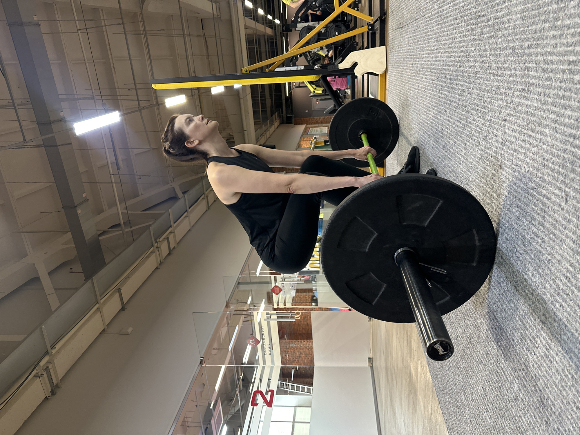 An athletic woman is performing a deadlift in a gym. She is wearing a black tank top and black leggings, with her hair pulled back in a ponytail. She is in a crouched position, with her hands gripping a barbell loaded with large black weight plates. The barbell has a green grip. The gym has a grey carpeted floor and exposed ceiling with industrial lighting and ventilation systems. In the background, there are other gym equipment and a brick wall visible through a glass partition. The gym appears to be a modern fitness facility. The gym is located in the city of Chaikovsky, Russia, as indicated by the Cyrillic text on the wall in the background which translates to "Fighter Club".