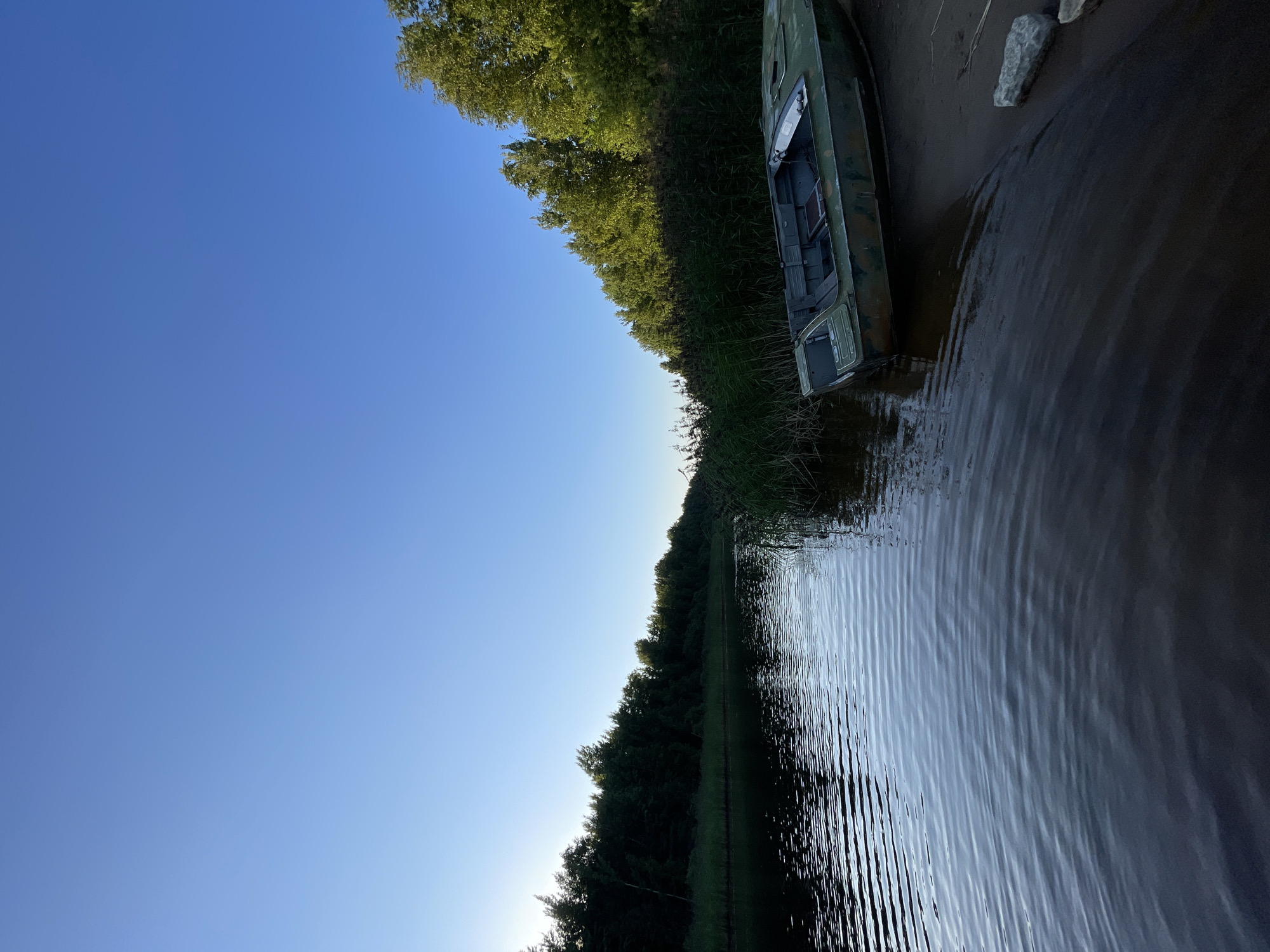 This is a vertical, eye-level, full shot of a river bank in Kobona, Russia. The sky is a clear, vibrant blue, taking up the top third of the image. On the right side, a line of green trees forms a backdrop against the sky. The river, in the foreground, is dark and calm, with gentle ripples distorting the reflection of the trees and the sky. On the sandy bank, a green, military-style boat is beached, facing right. The boat is empty and appears to be made of metal, with camouflage markings. There are a few rocks scattered on the bank near the boat. The overall scene is serene and depicts a quiet, natural landscape.