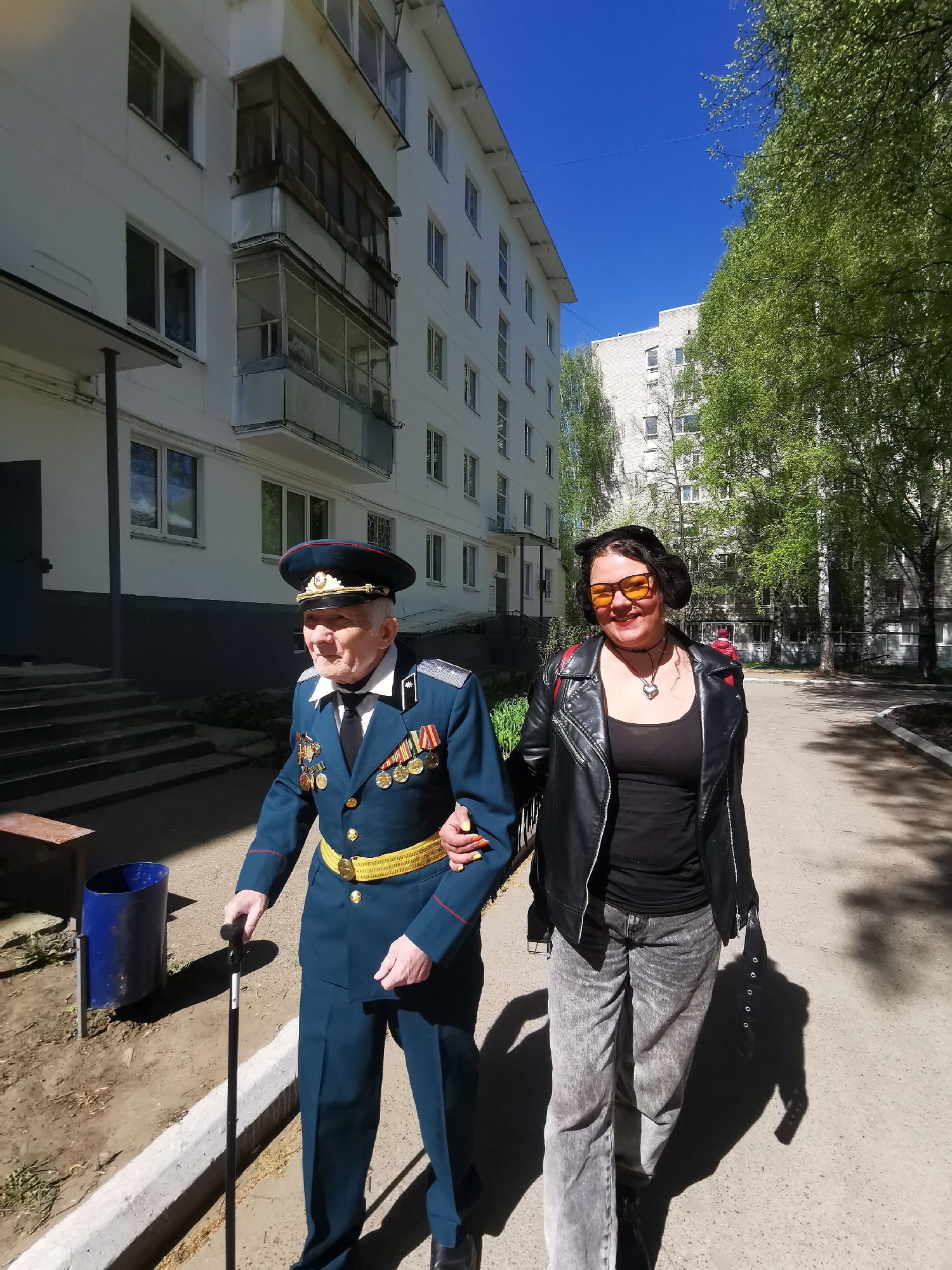 An elderly man in a military uniform walks arm-in-arm with a younger woman on a sunny day. The man is wearing a blue military uniform with gold trim and numerous medals. He is using a black cane. The woman is wearing a black leather jacket, a black top, and grey jeans. She has short dark hair and is wearing orange sunglasses. They are walking on a paved path next to an apartment building. The building is white with multiple windows and a balcony. In the background, there are trees with green leaves and another apartment building. The scene is in the city of Chaikovsky, Russia.