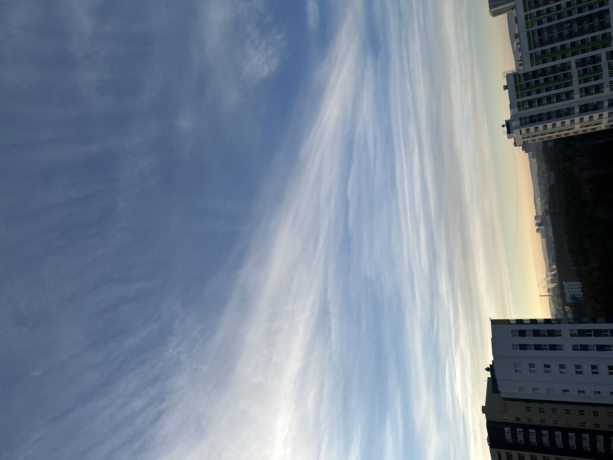 The image depicts a view of the sky with cirrus clouds over Yekaterinburg, Russia. Two tall apartment buildings are visible on the right side of the frame. In the distance between the buildings, there is a horizon showing signs of early morning or late evening light with a light yellow-orange tint. A darker, wooded area is partially obscured by the buildings.