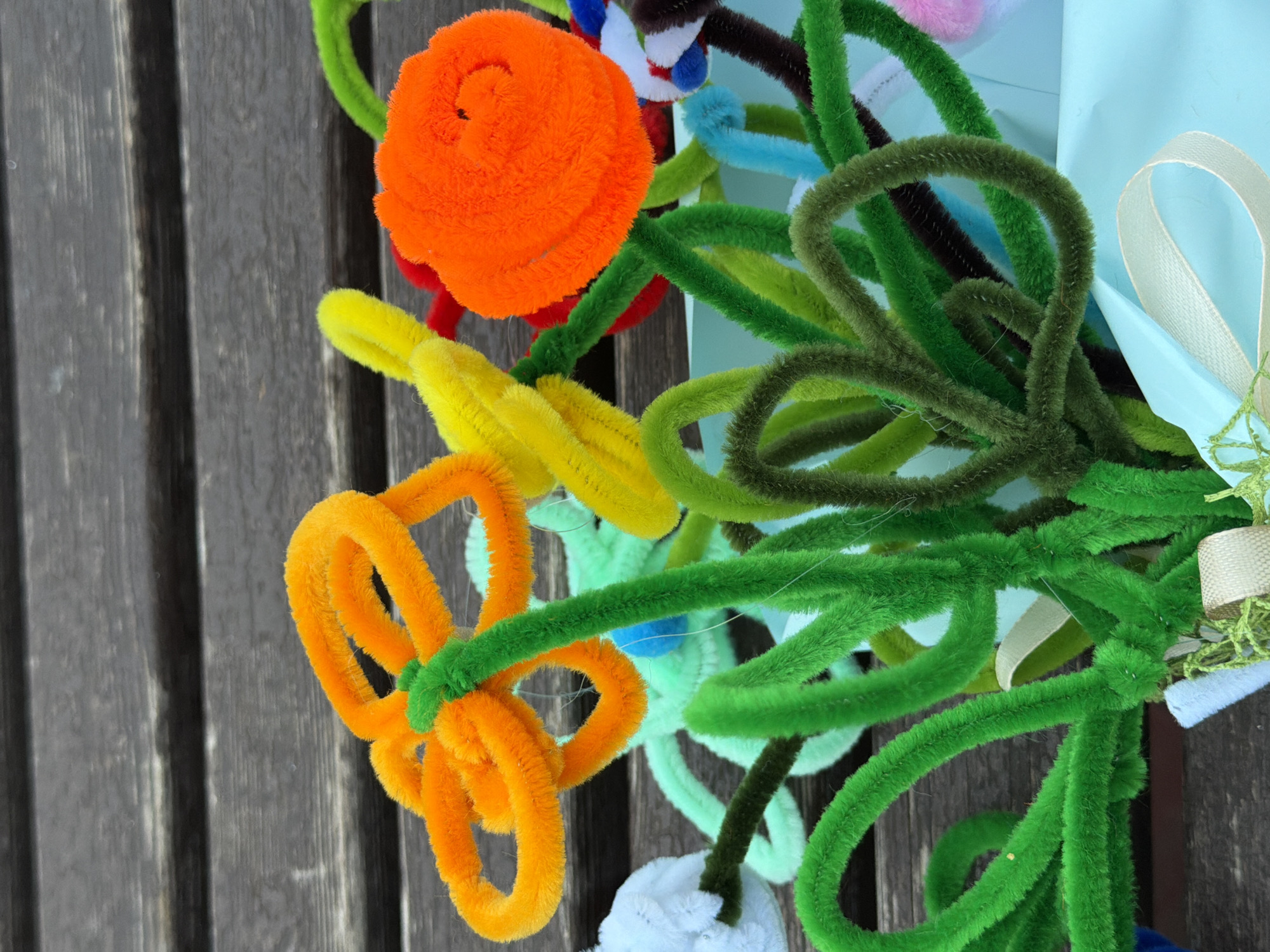 The image shows a close-up of a bouquet of handmade pipe cleaner flowers arranged in a light blue container. The flowers are in various colors, including orange, yellow, green, and white, with some darker green and brown stems also visible. The arrangement is set against a backdrop of weathered dark wood planks. The lighting is natural, casting soft shadows on the flowers and the wooden surface. The image is focused on the flowers, with the background slightly out of focus, highlighting the texture and detail of the pipe cleaner creations. No people or specific city location are visible.
